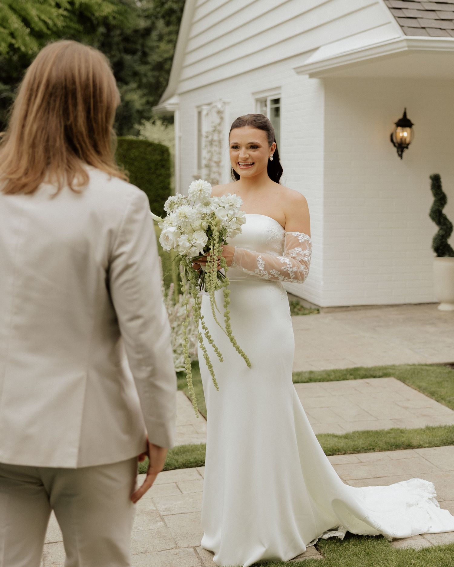 Bride in white off-shoulder gown holding cascading white bouquet during first look at elegant outdoor wedding venue.