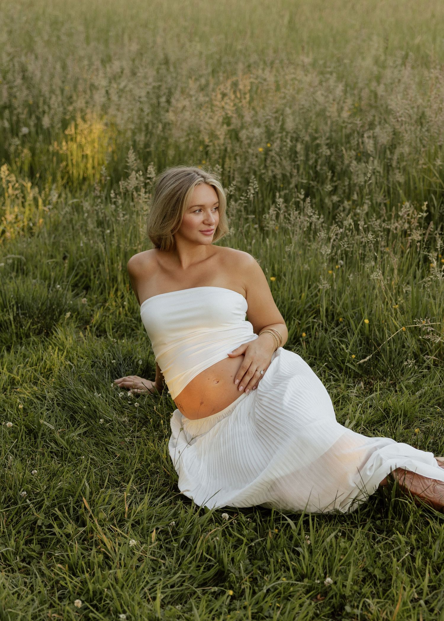 Pregnant woman in white tube top and skirt sitting in a lush green field during golden hour.