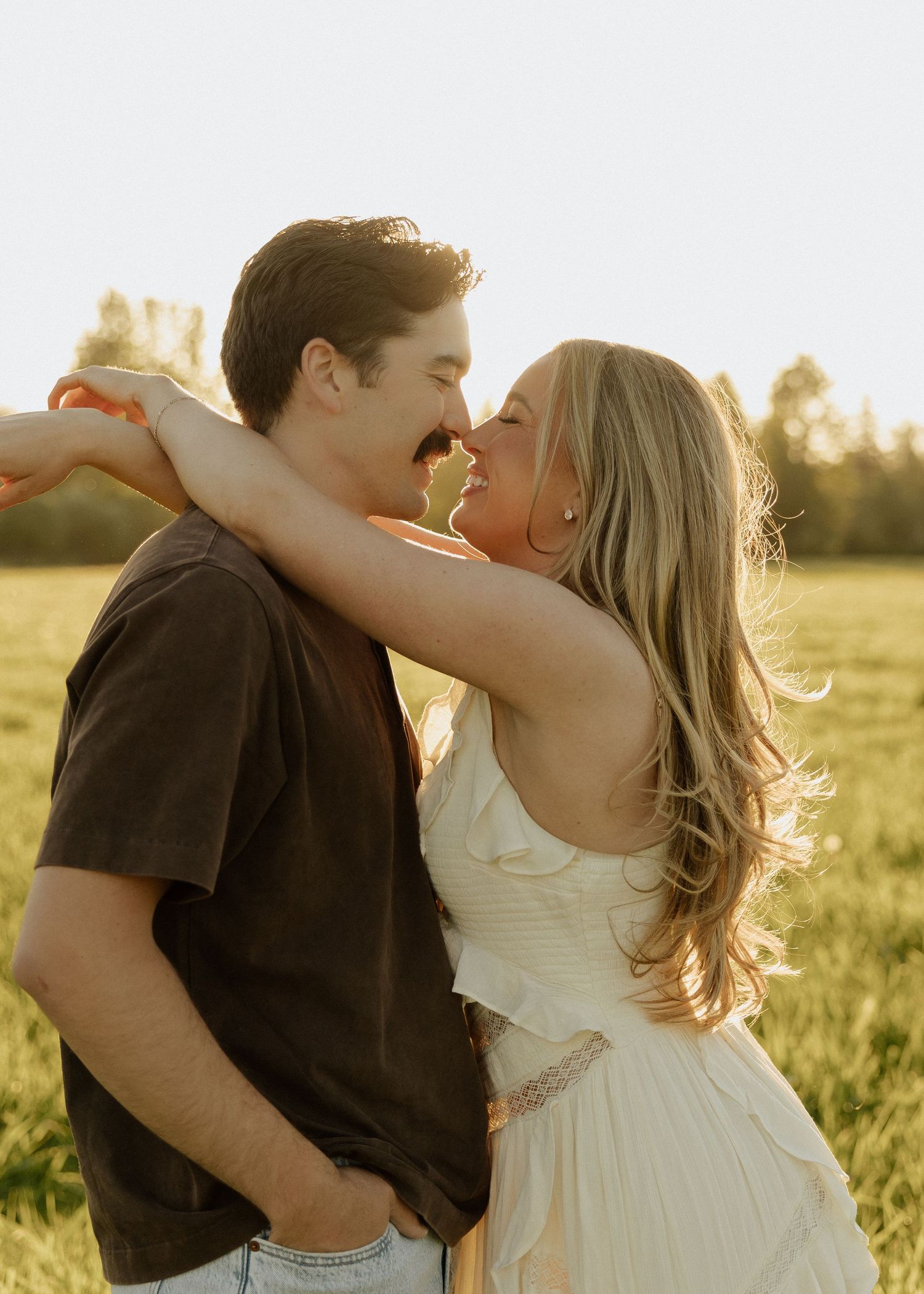 A couple shares a romantic almost-kiss in a sunlit field, the woman in a white dress with arms around the man.