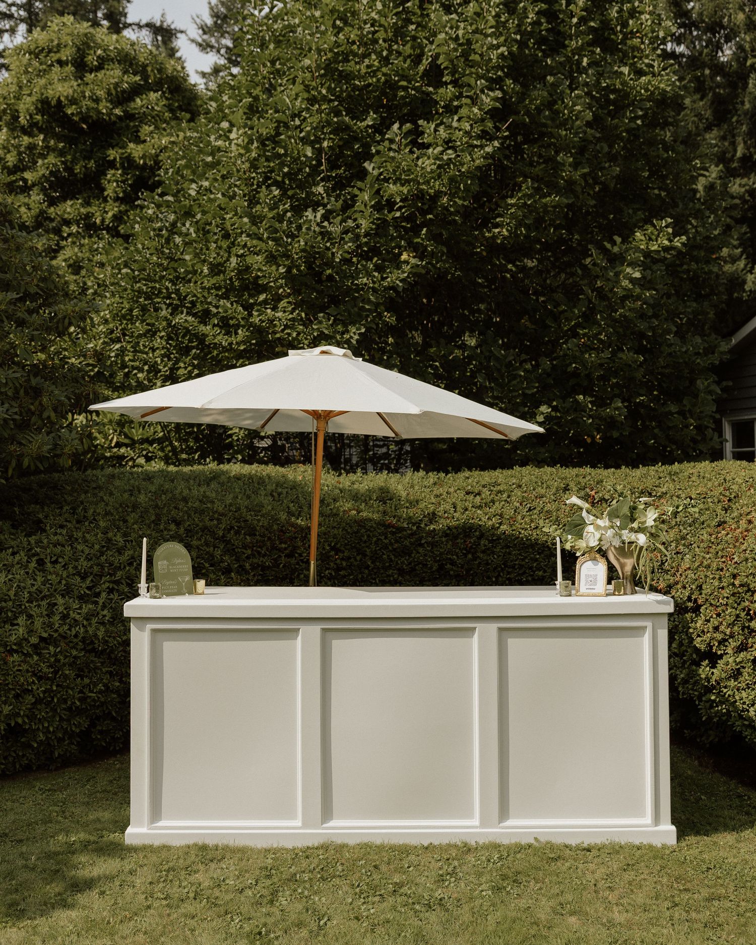 White outdoor bar setup with patio umbrella surrounded by lush green hedges and trees at a garden wedding.