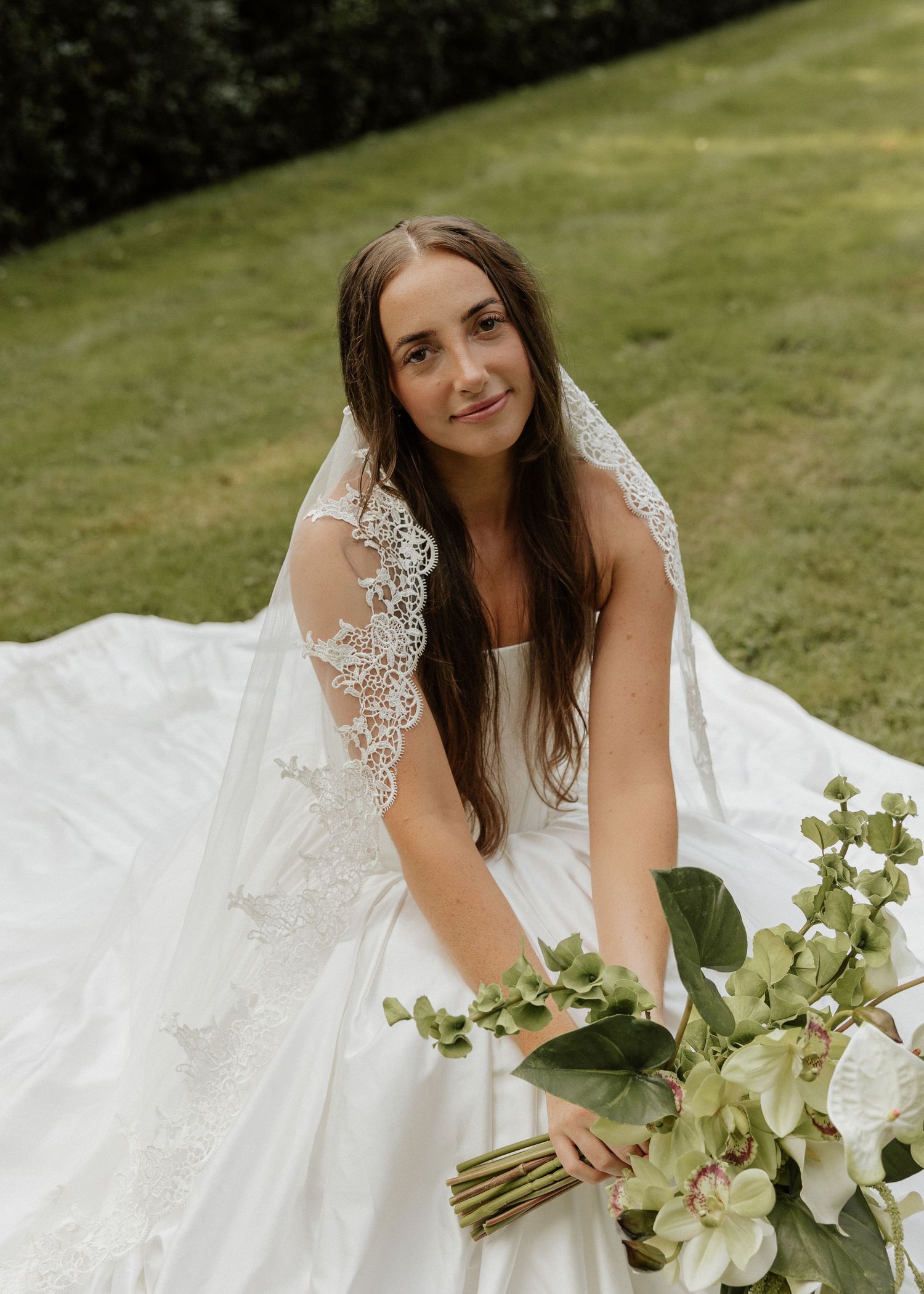 Bride in lace veil and white gown holding green orchid bouquet outdoors.