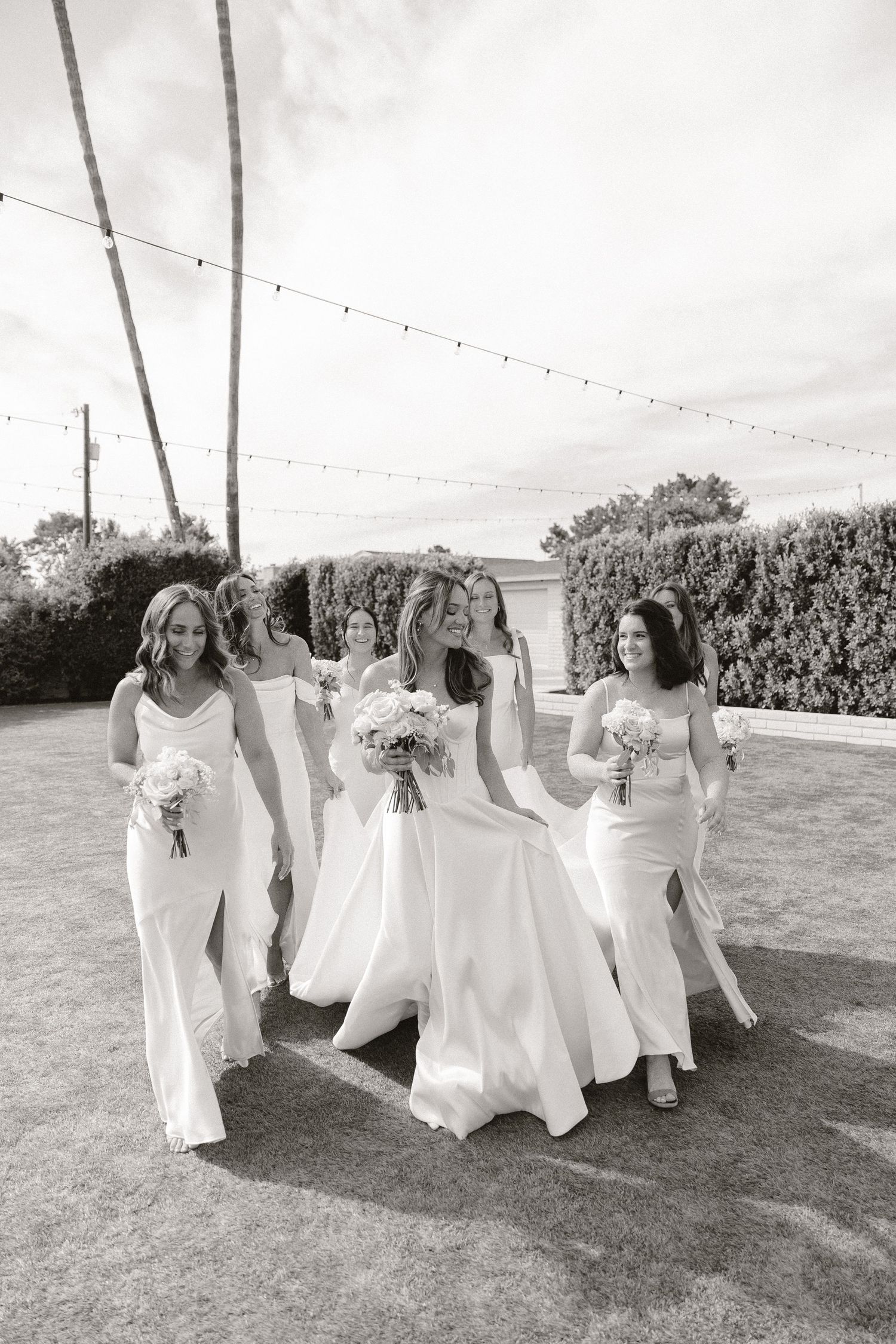 Bride and bridesmaids in white gowns walking outdoors with bouquets under string lights in black and white photo.