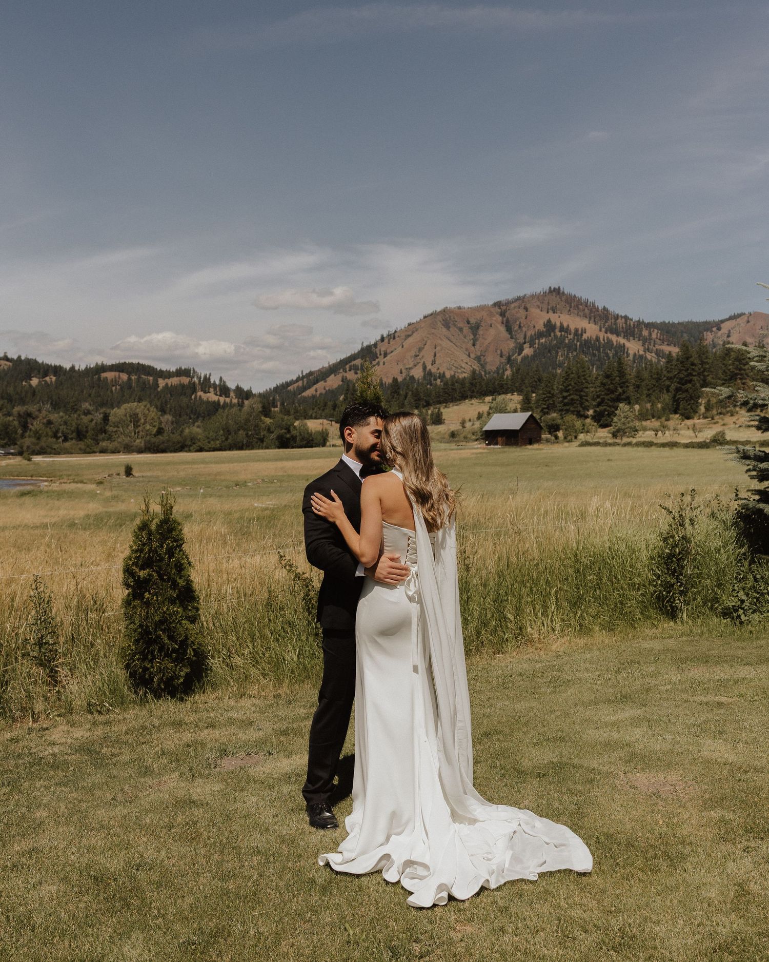 A bride and groom share a romantic embrace in front of a stunning mountain landscape and golden meadow.