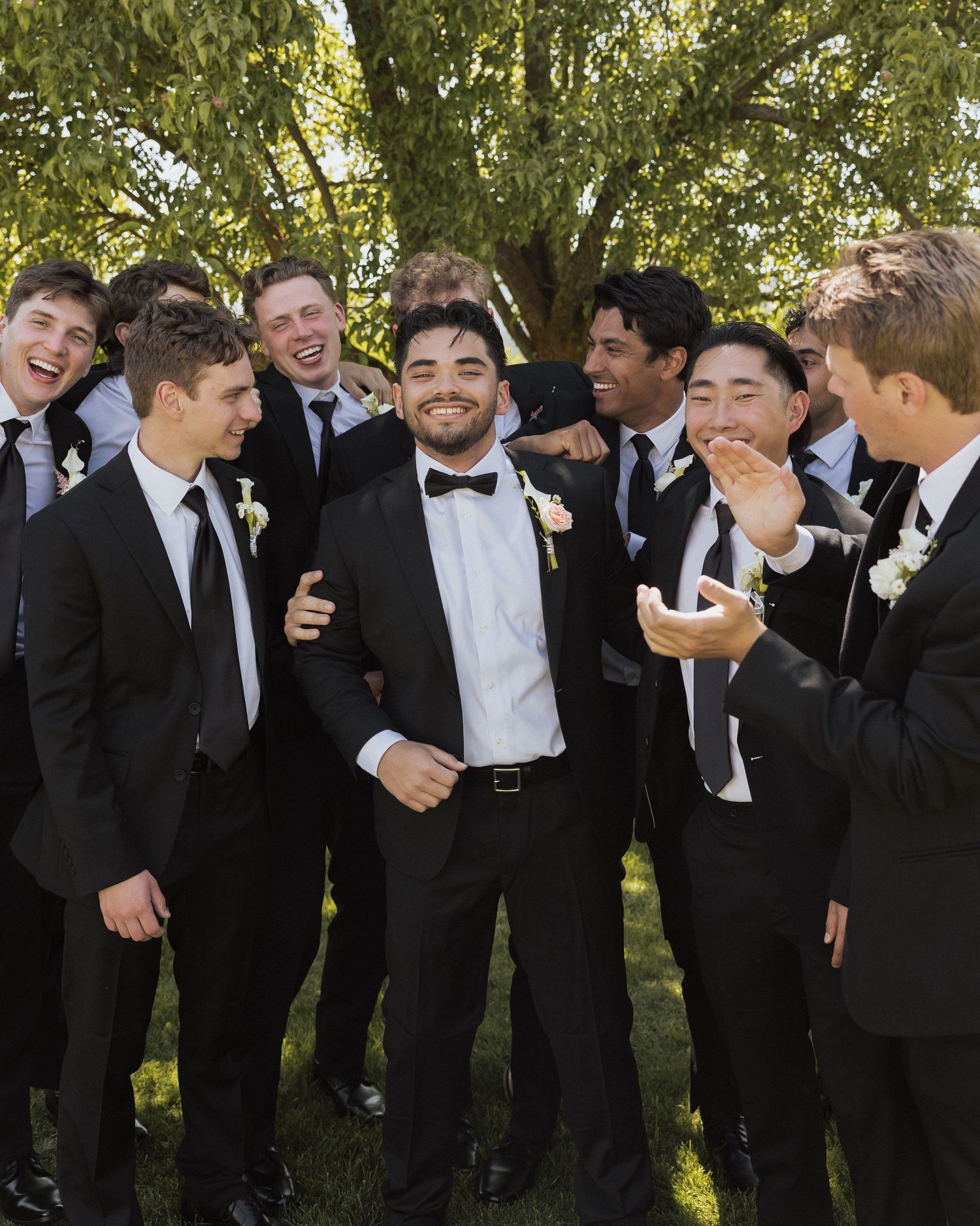 Groom in black tuxedo laughing with groomsmen in matching suits outdoors at a wedding.
