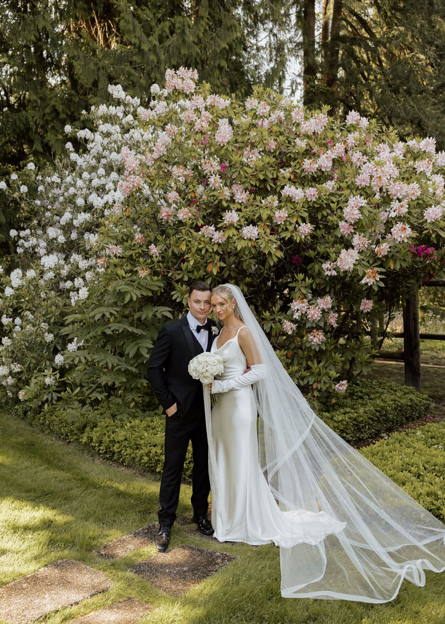 Bride and groom pose in lush garden with blooming rhododendrons, bride wearing white gown and veil.