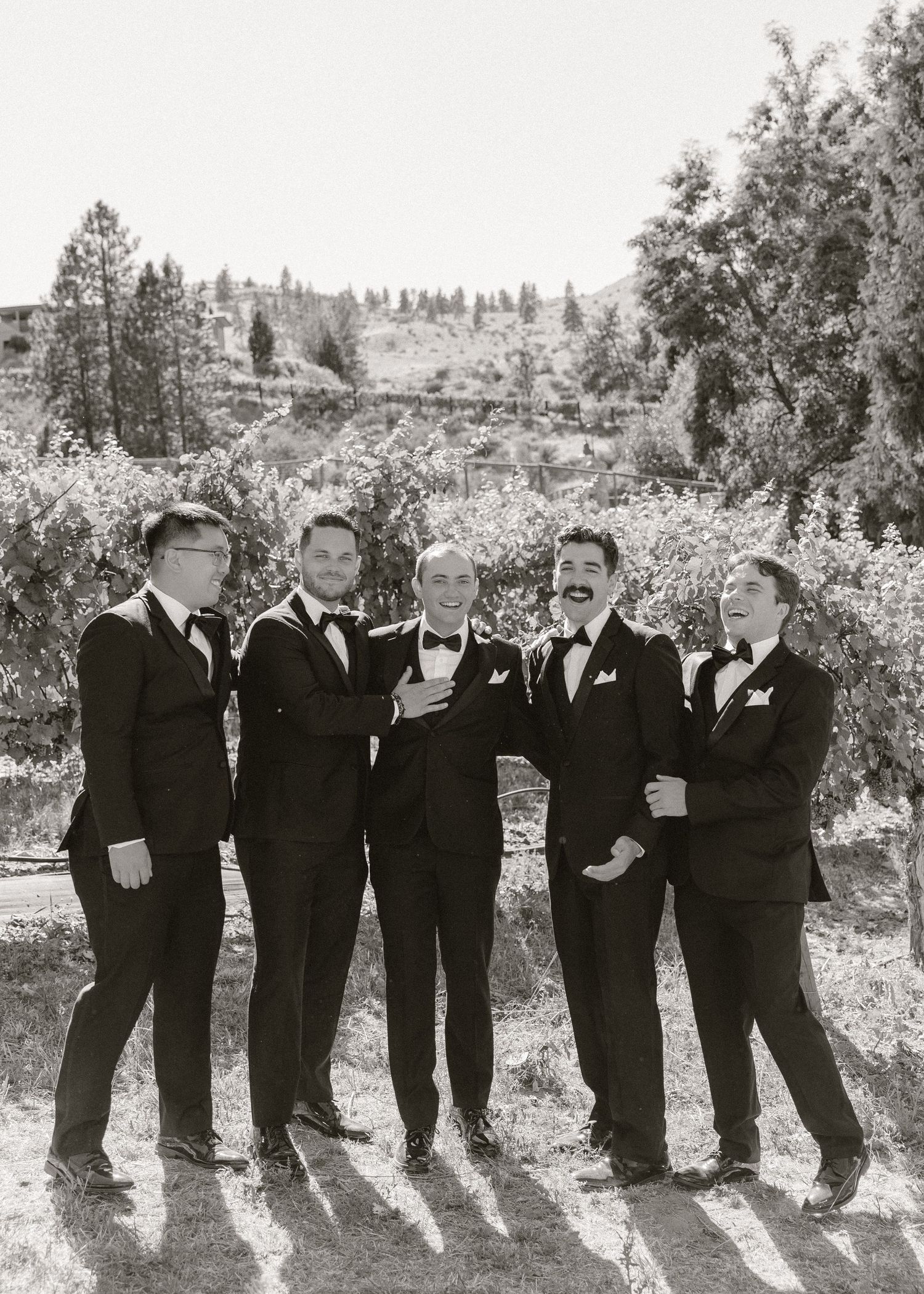 Five groomsmen in black tuxedos and bow ties pose together outdoors with trees and hills in black and white photo.
