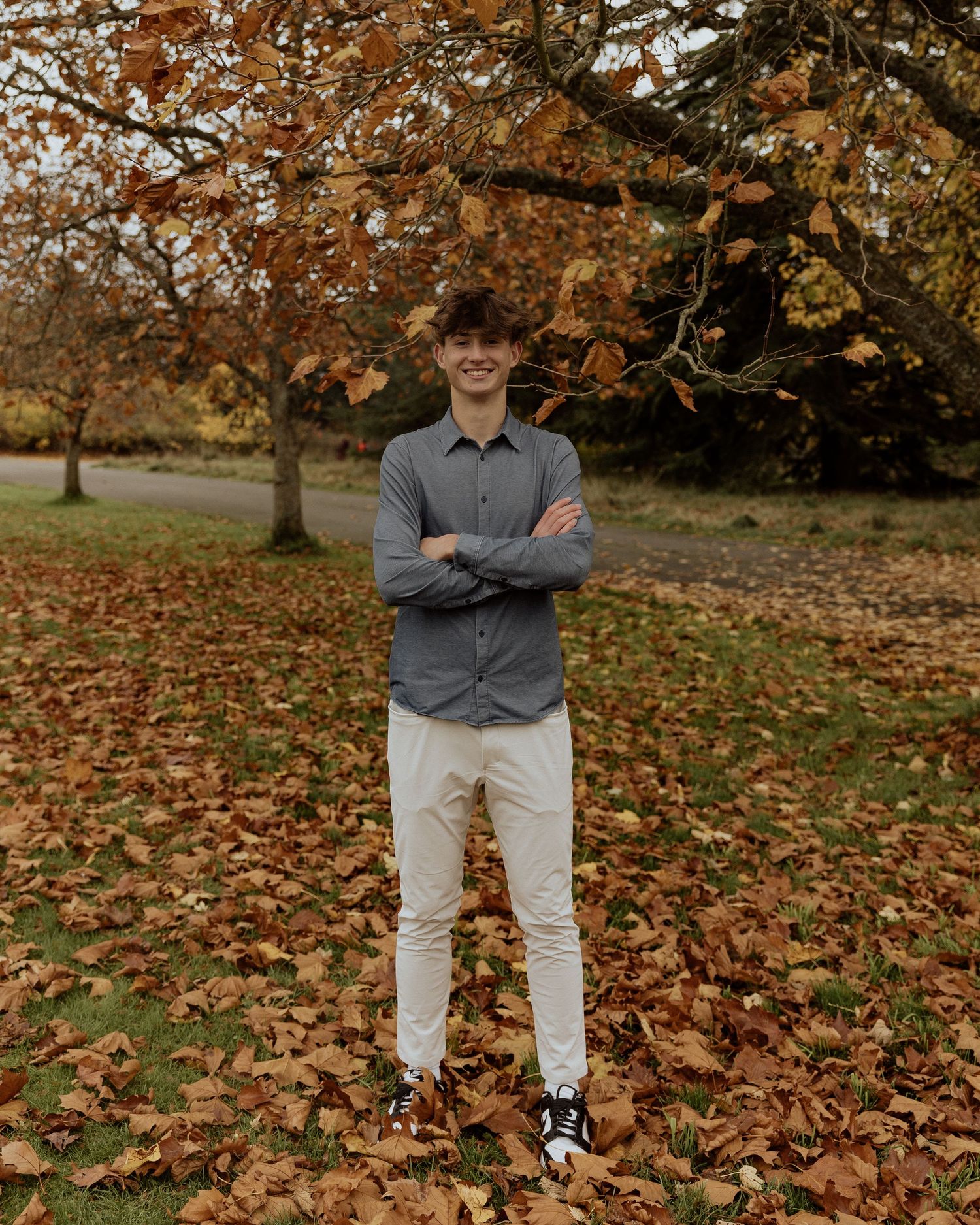 A person in grey shirt and white pants stands smiling among fallen autumn leaves.