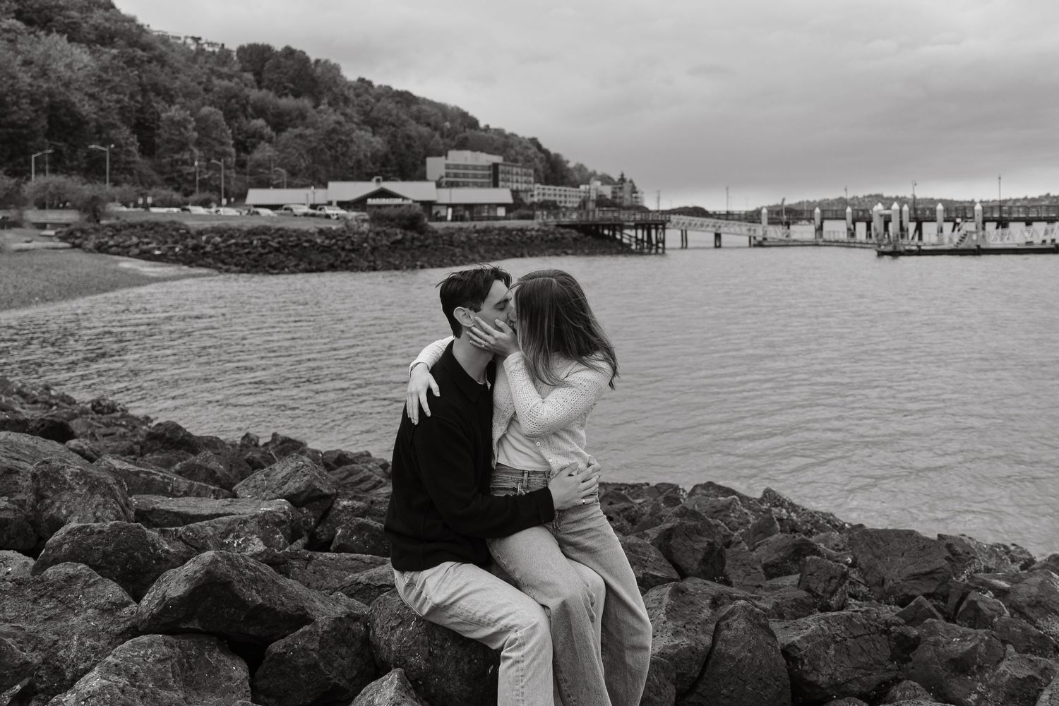 A couple sharing a romantic moment on rocky shoreline with harbor view.