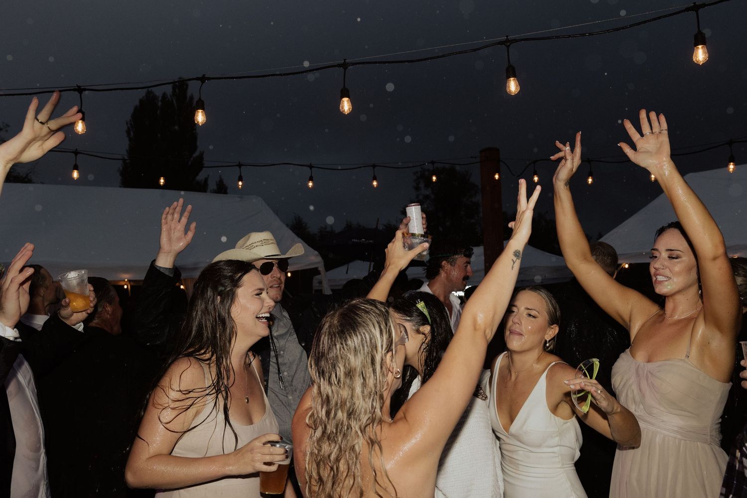 People dancing with raised arms under string lights at an outdoor party.