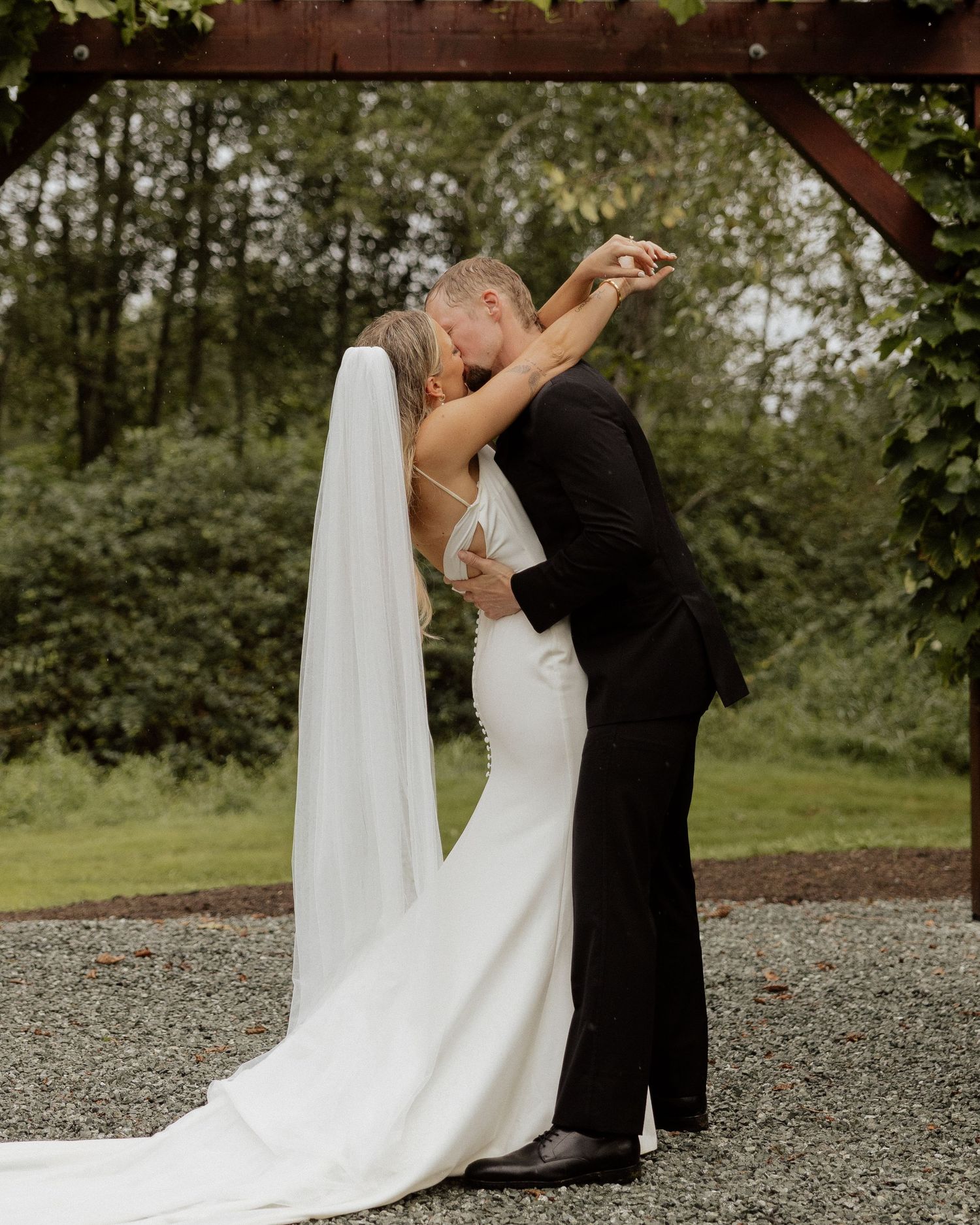 A romantic embrace under a rustic wooden pergola decorated with greenery.