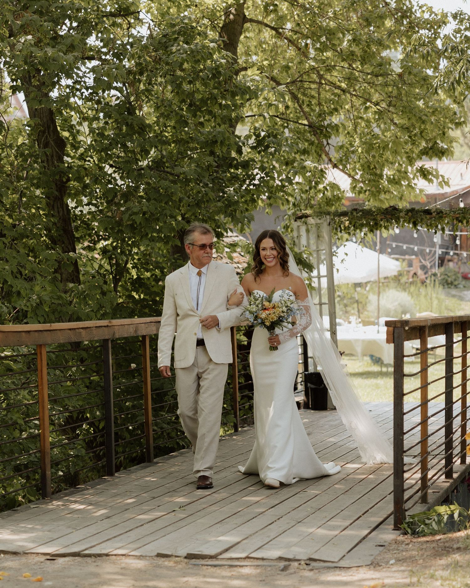Wedding procession on wooden bridge with couple walking arm in arm under green trees.