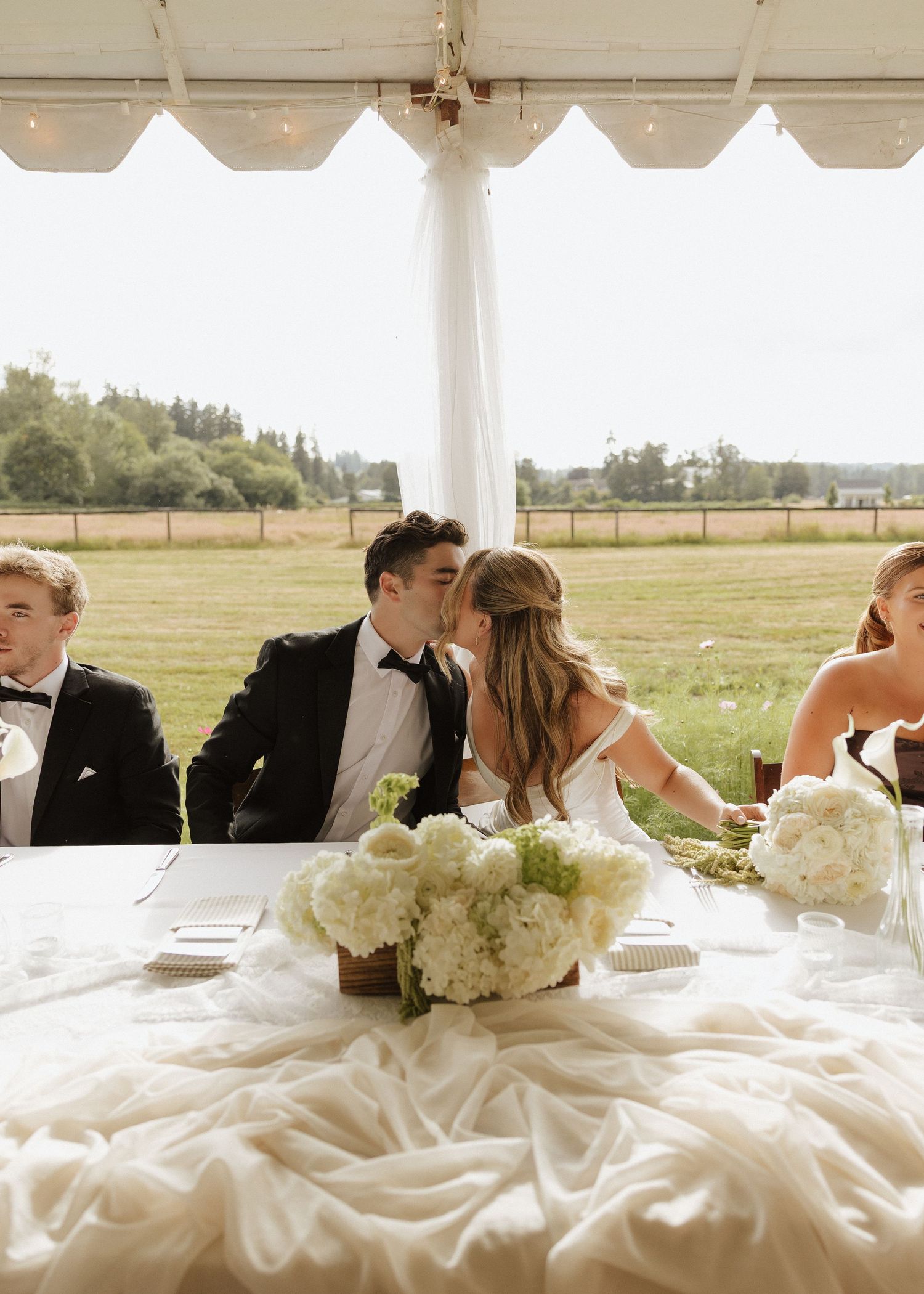 Bride and groom share a kiss at elegant outdoor wedding reception under white tent with hydrangea centerpieces.