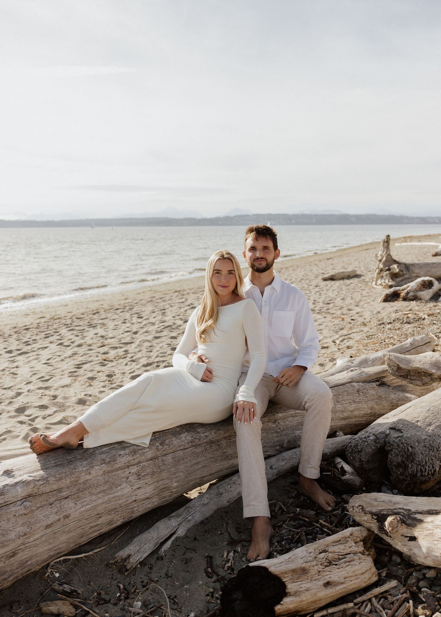 Couple in white outfits sitting on driftwood log on sandy beach with ocean backdrop.