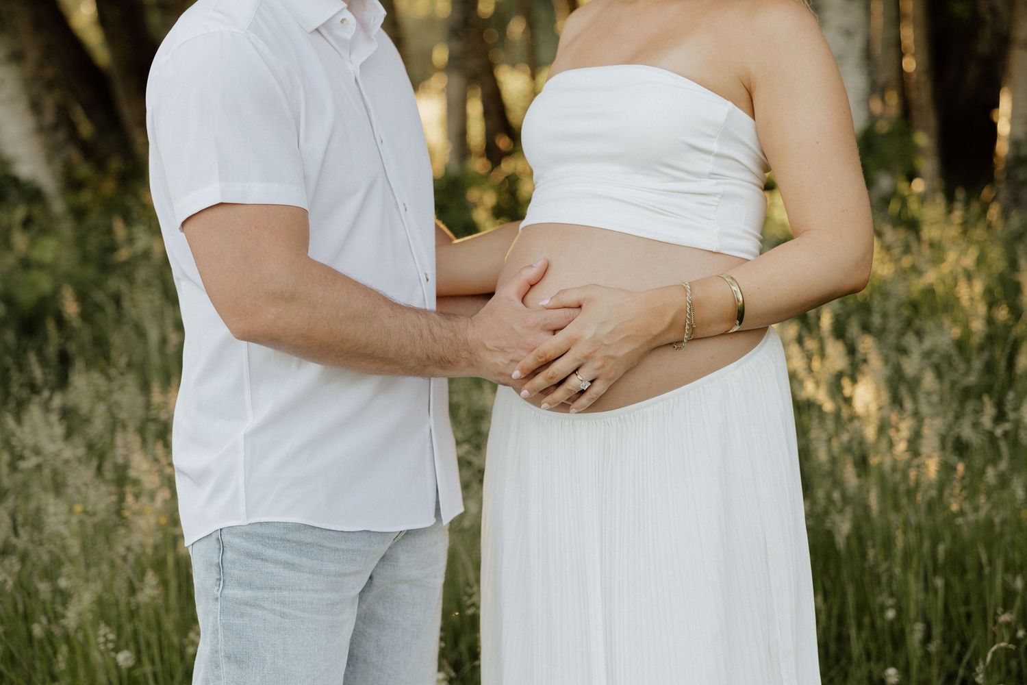 Couple in white outfits holding pregnant belly outdoors in sunlit forest setting.