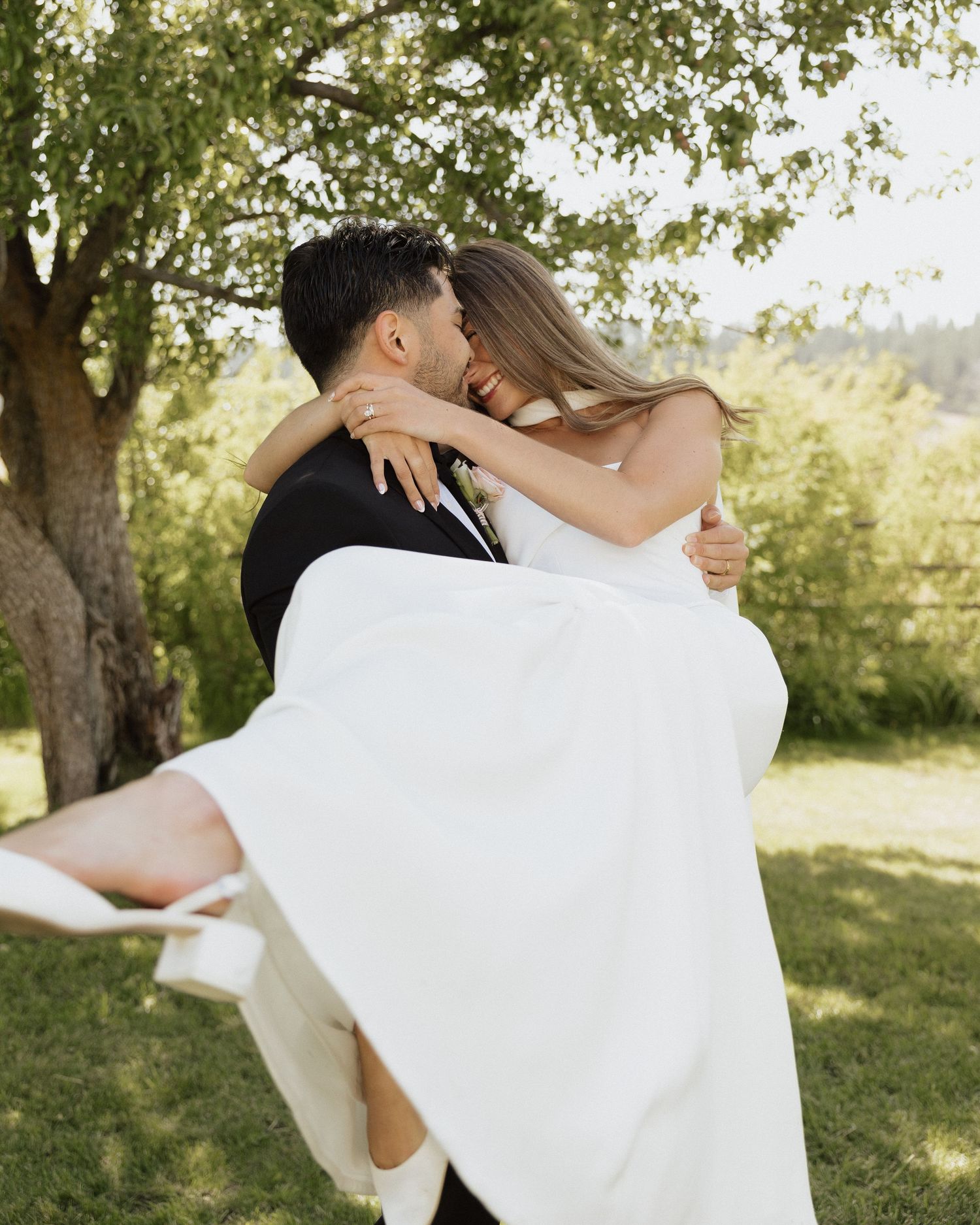 Groom lifts bride in white wedding dress outdoors, sharing a romantic kiss under a lush green tree.
