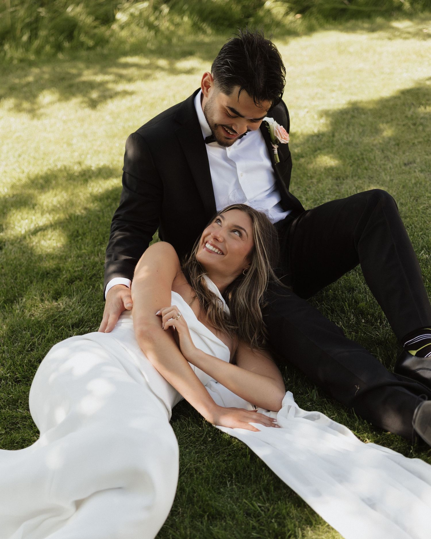 Bride in white gown reclines on grass smiling up at groom in black suit during outdoor wedding photos.