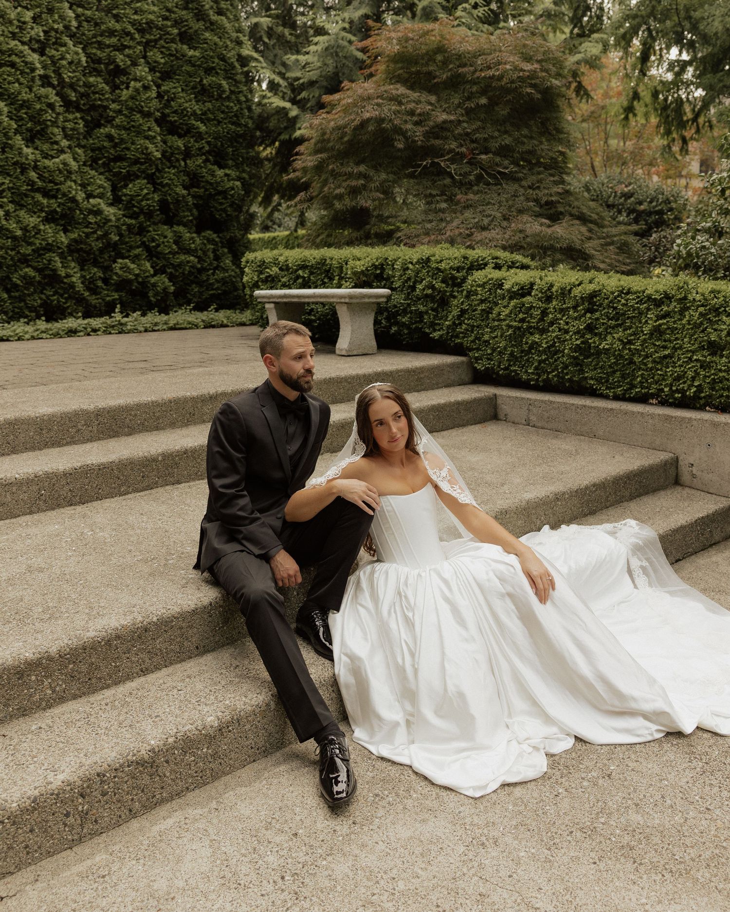 Bride in off-shoulder white gown and groom in black suit sitting on stone steps in a lush garden setting.