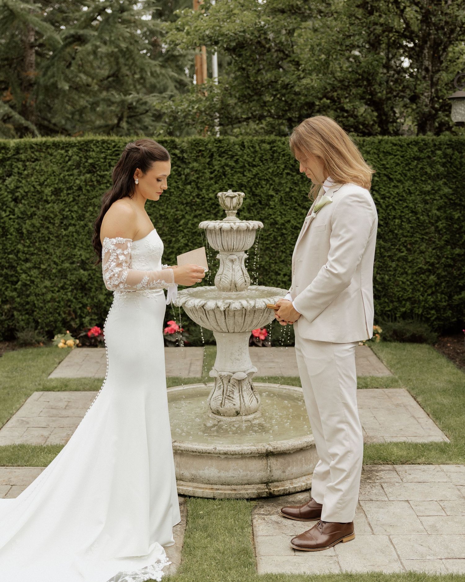 Bride and groom exchange vows beside a stone fountain in a lush garden during an outdoor wedding ceremony.