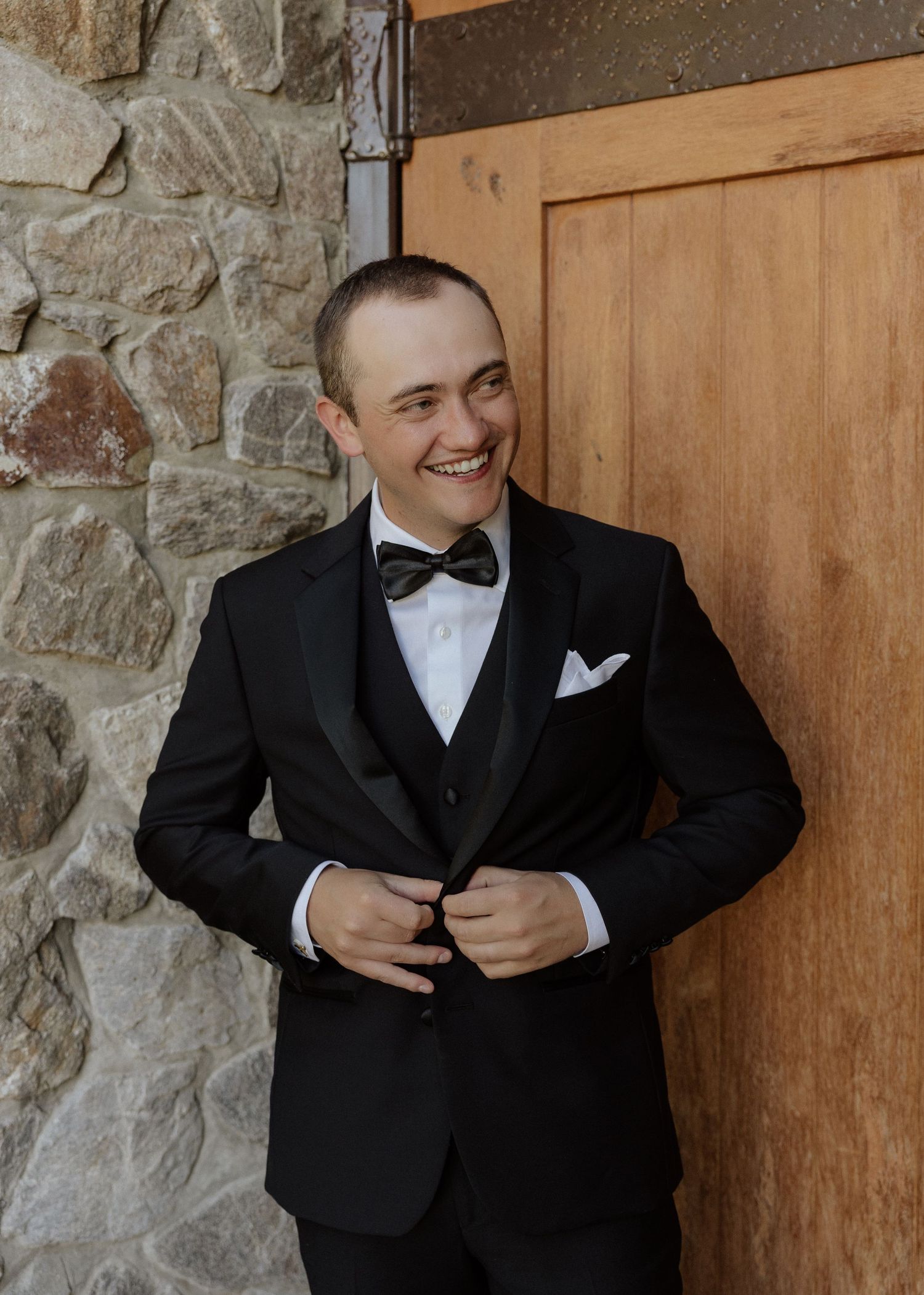 Smiling groom in black tuxedo with bow tie and white pocket square, standing by wooden door and stone wall.