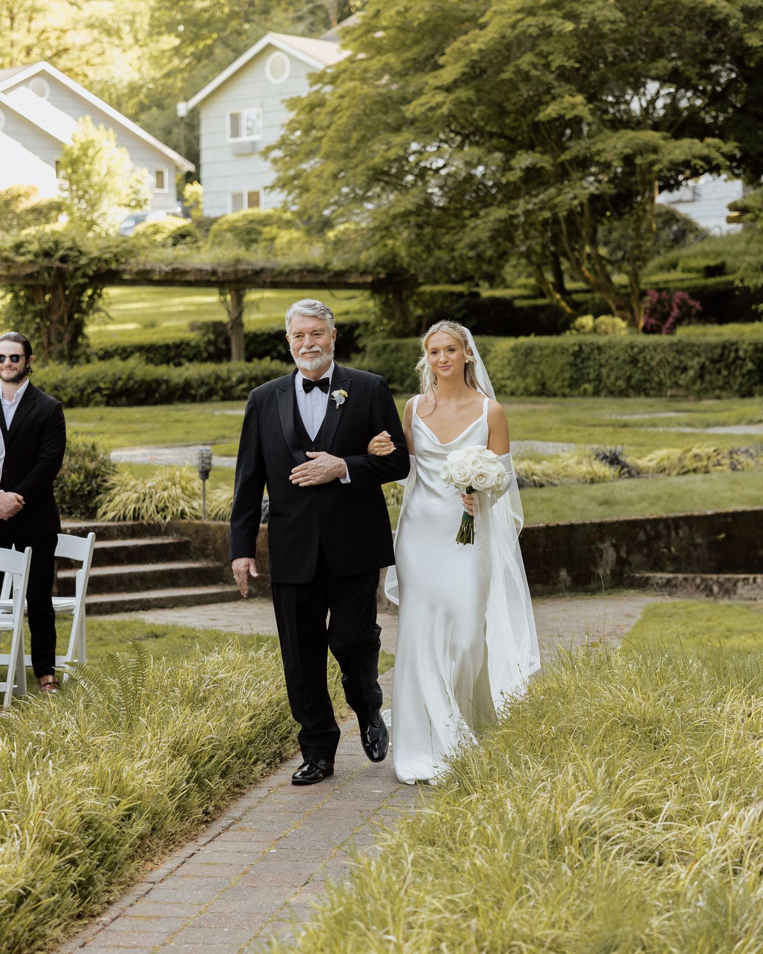 Bride in white gown walks down outdoor aisle with older gentleman in tuxedo at garden wedding ceremony.