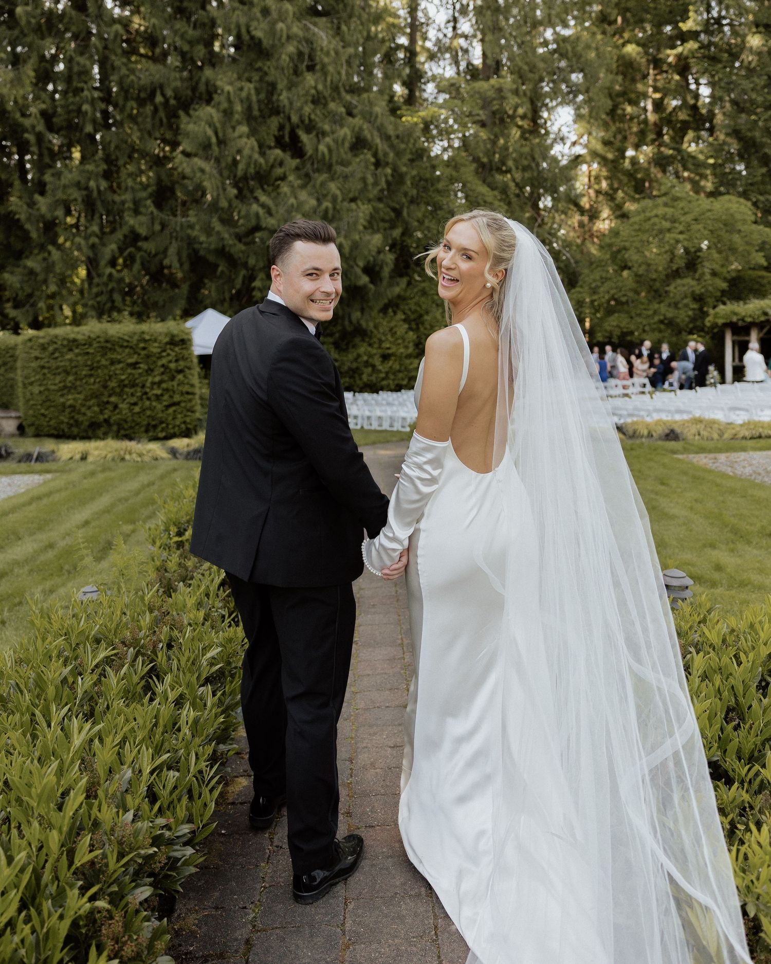 A bride in a white gown and veil holds hands with a groom in a black tuxedo as they walk through a garden.