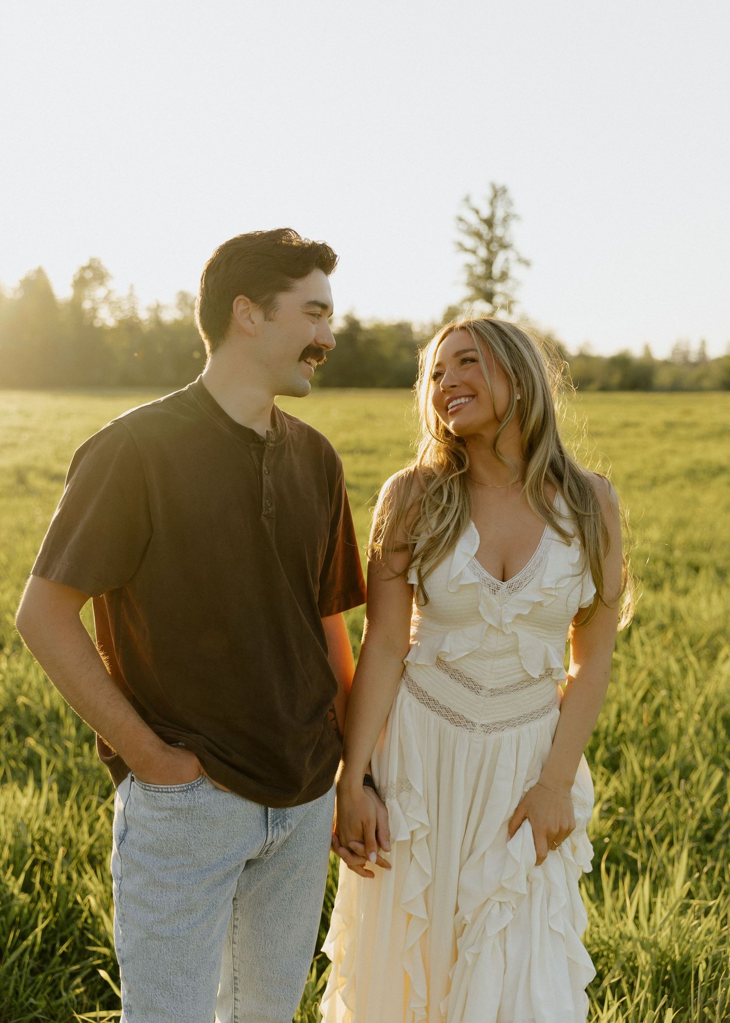 A happy couple holds hands walking through a sunlit green field during golden hour outdoors.