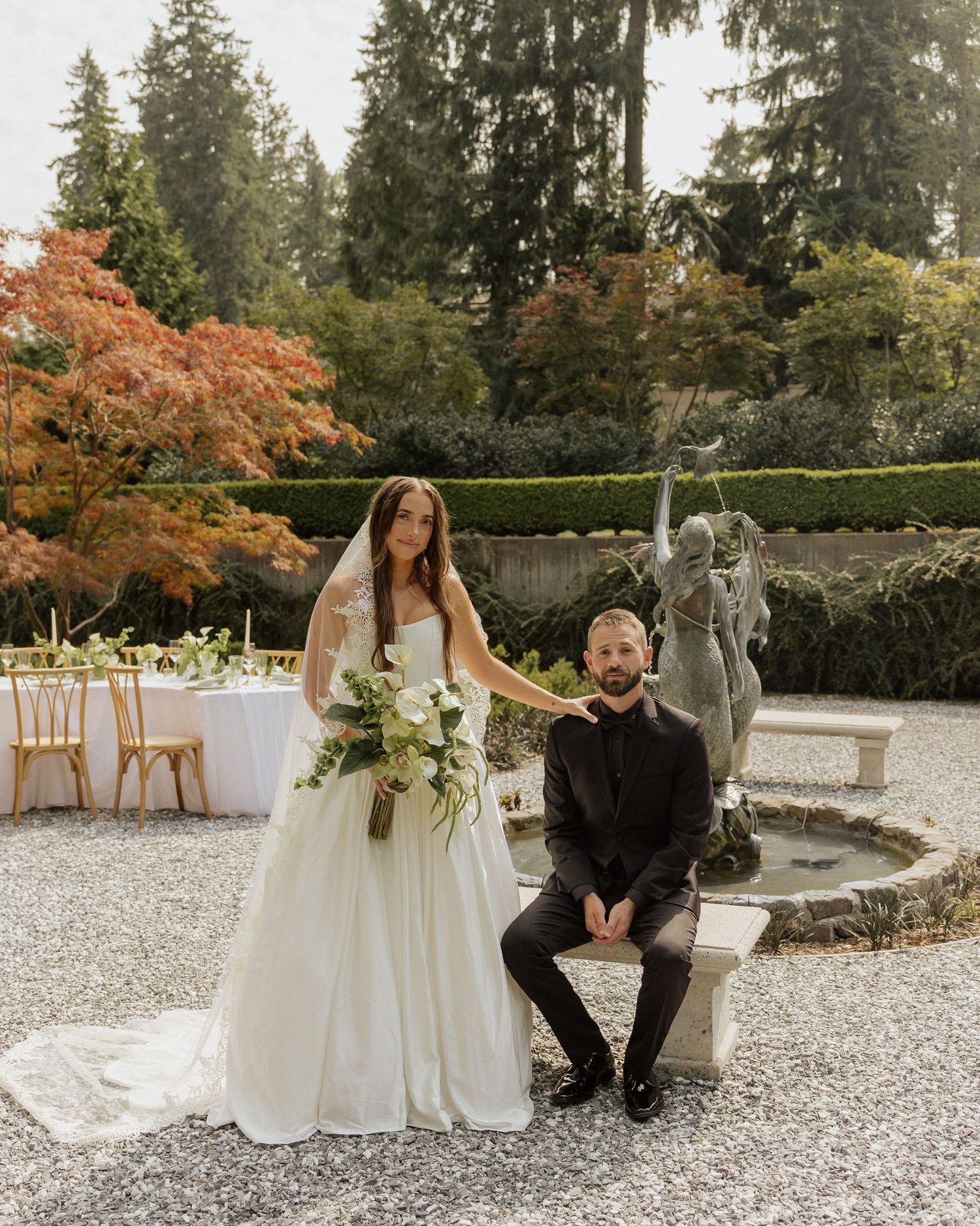 Bride in white gown with bouquet stands beside groom in black suit near stone fountain in autumn garden.