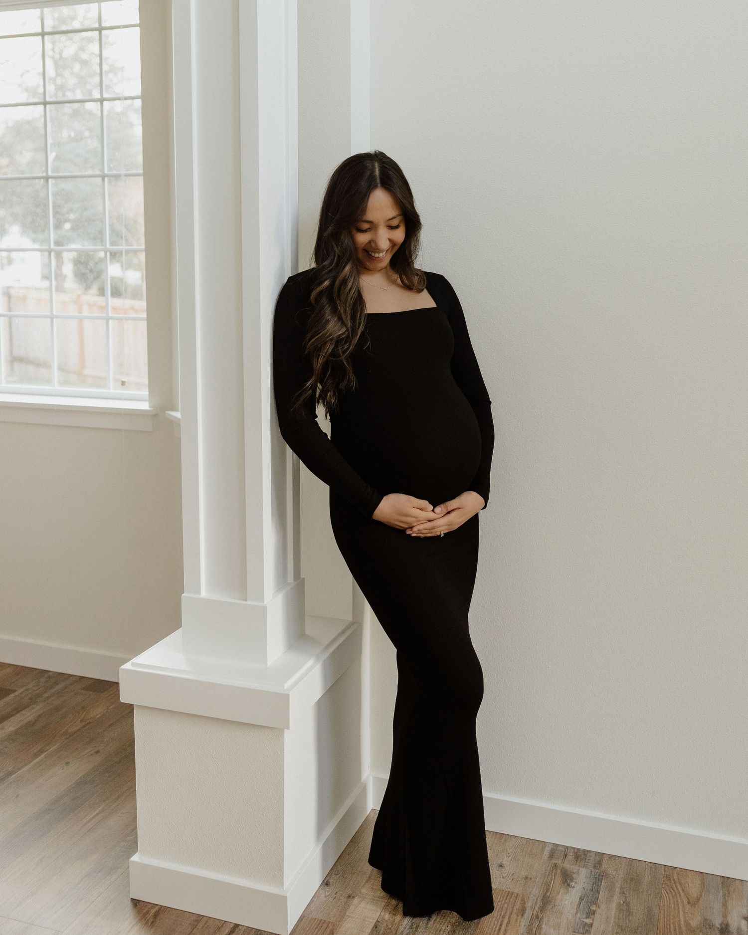 Pregnant woman in black dress stands by a window column in a minimalist setting.