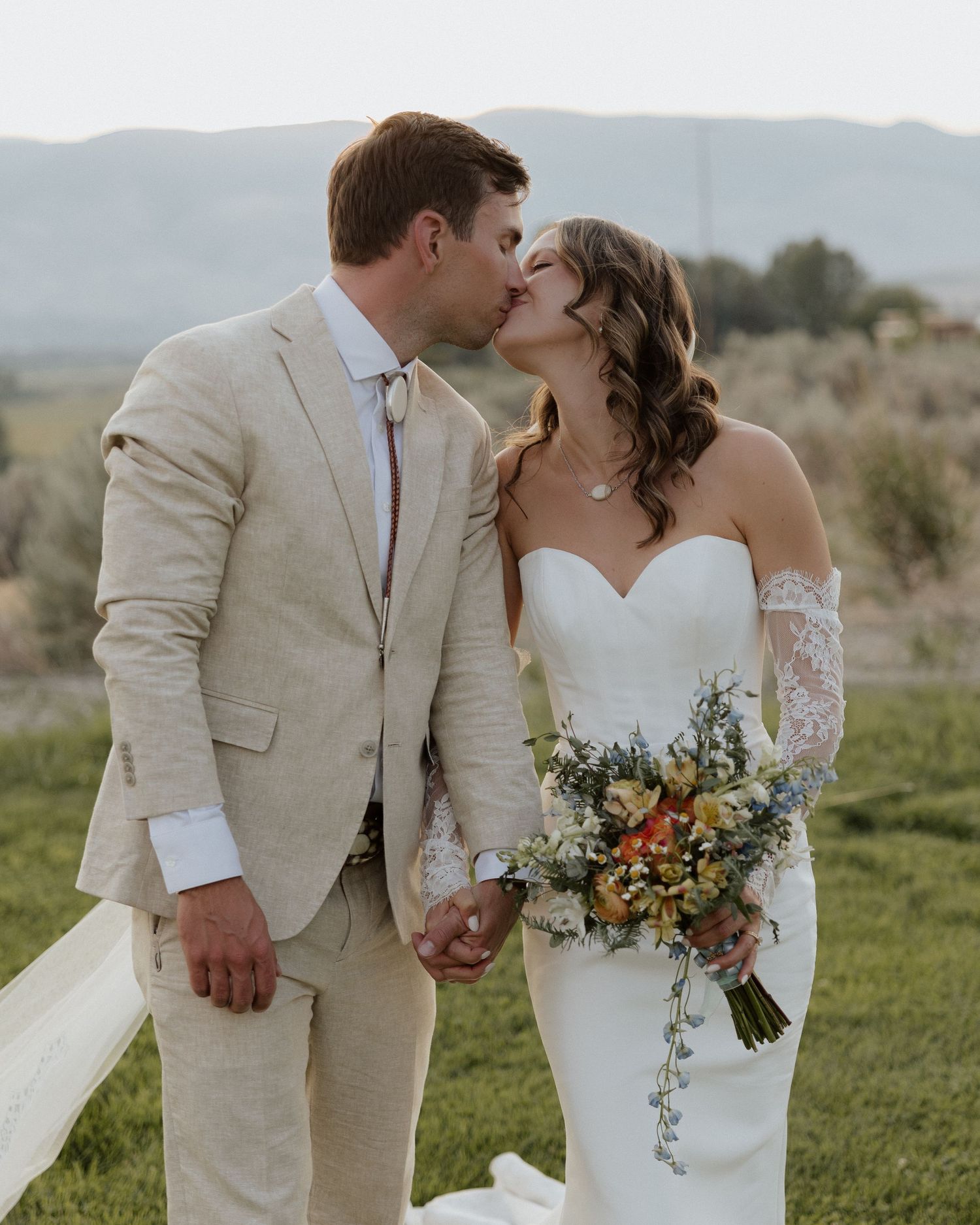 Bride in lace sleeve gown kisses groom in beige suit while holding vibrant bouquet.