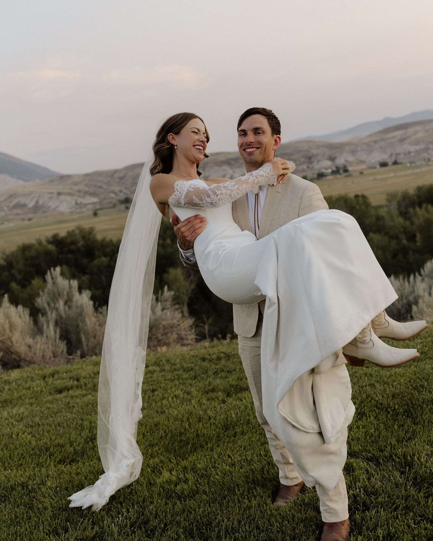 Groom carries laughing bride in white dress against mountainous backdrop during sunset photoshoot.