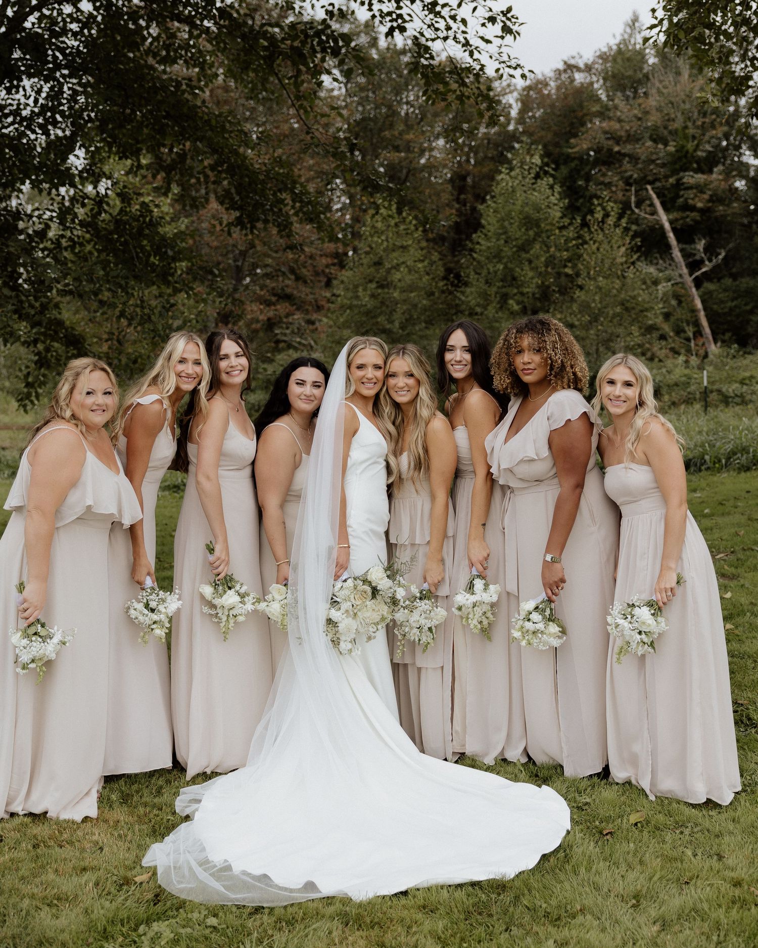 Bridesmaids in champagne-colored dresses pose with bride in flowing white gown for formal wedding photo.