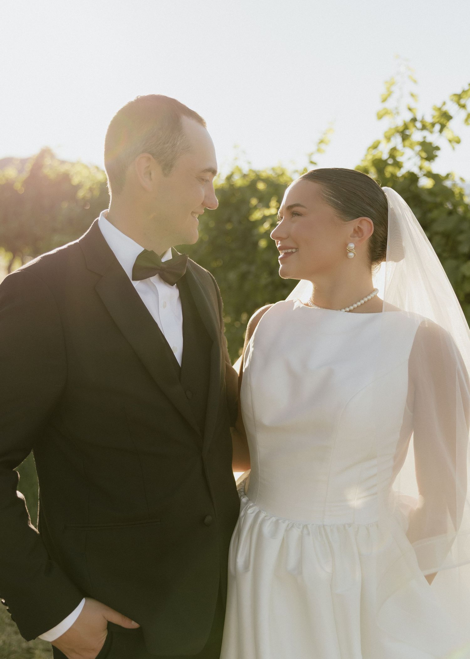 Bride in elegant white gown and groom in black tuxedo share a joyful glance in golden sunlight.