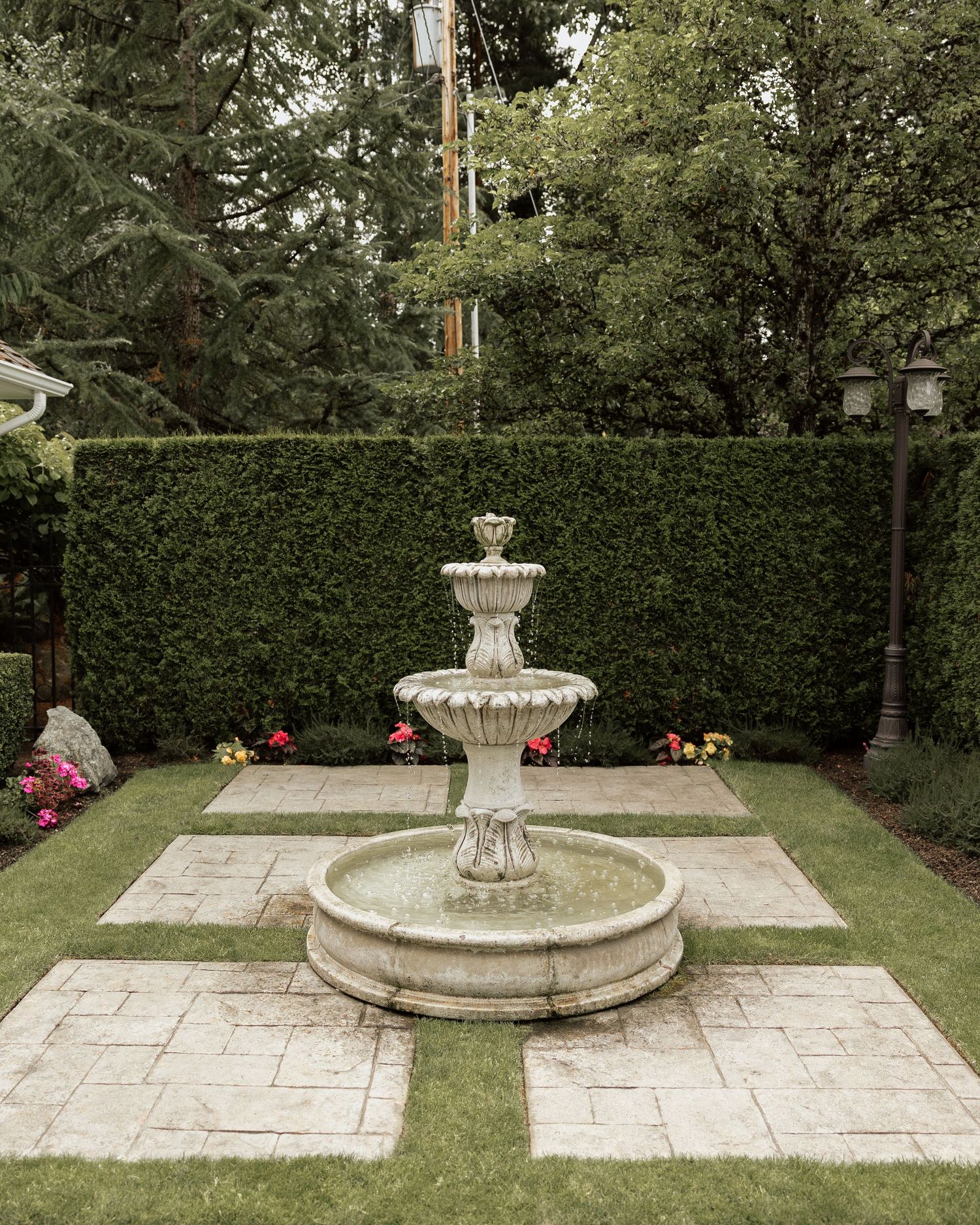 Elegant three-tier stone fountain centerpiece in manicured garden with hedgerow backdrop.