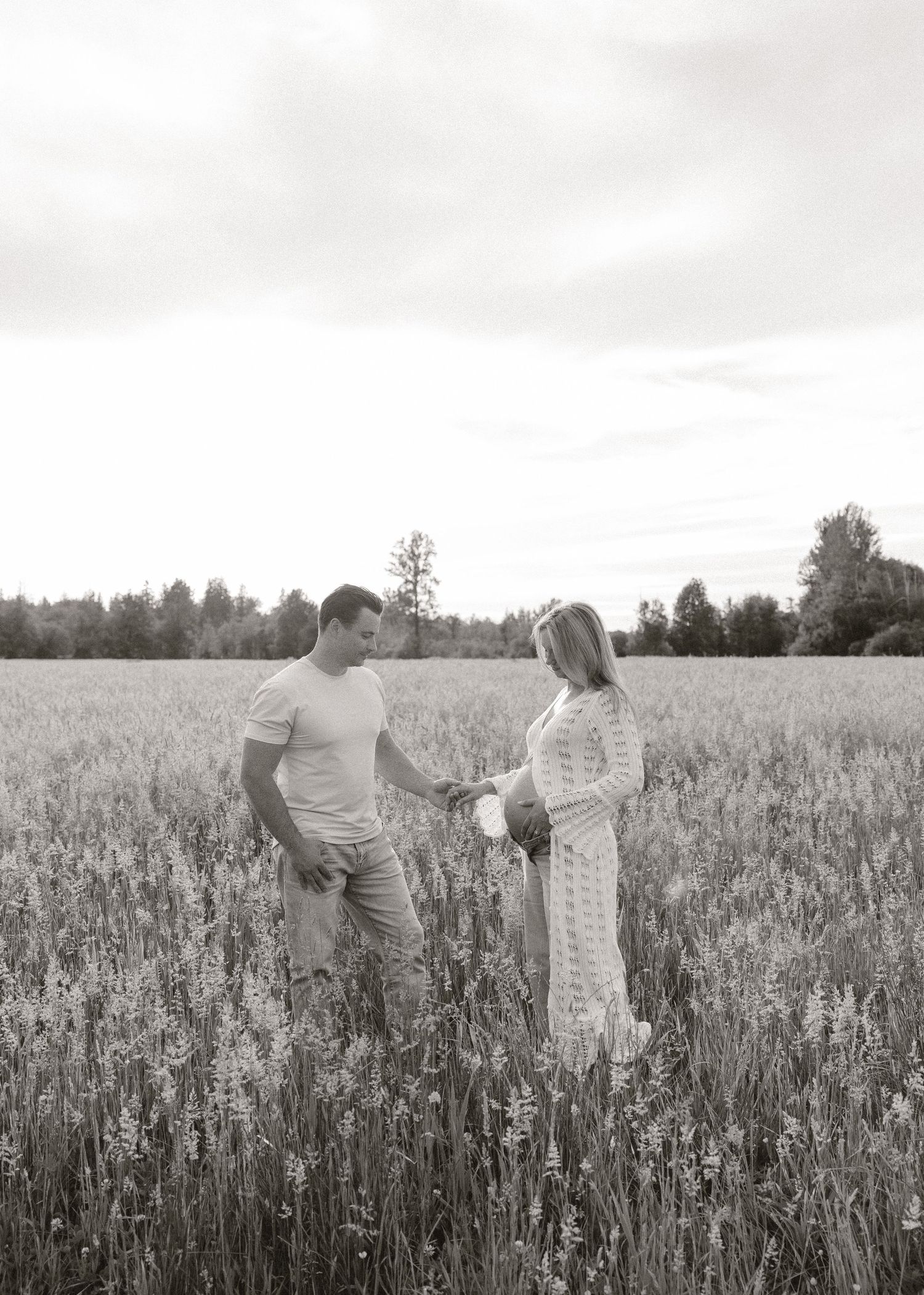 Black and white photo of a couple standing in a field of tall grass, facing each other, with trees in the background.