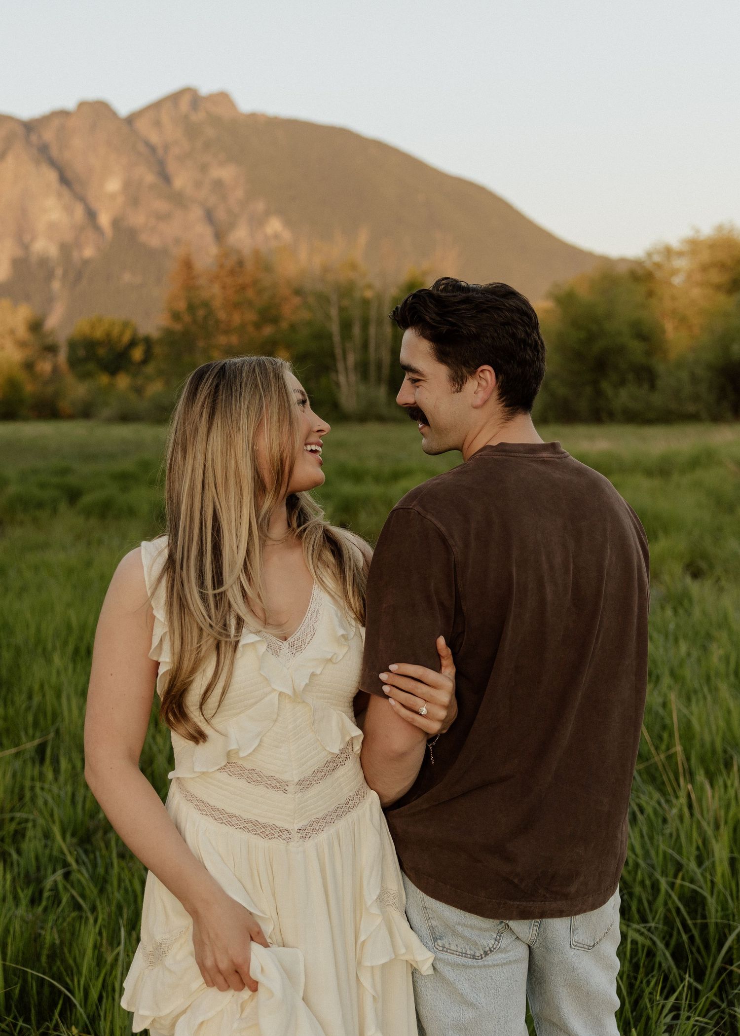 A laughing couple embraces in a green meadow with a stunning mountain range glowing at sunset.