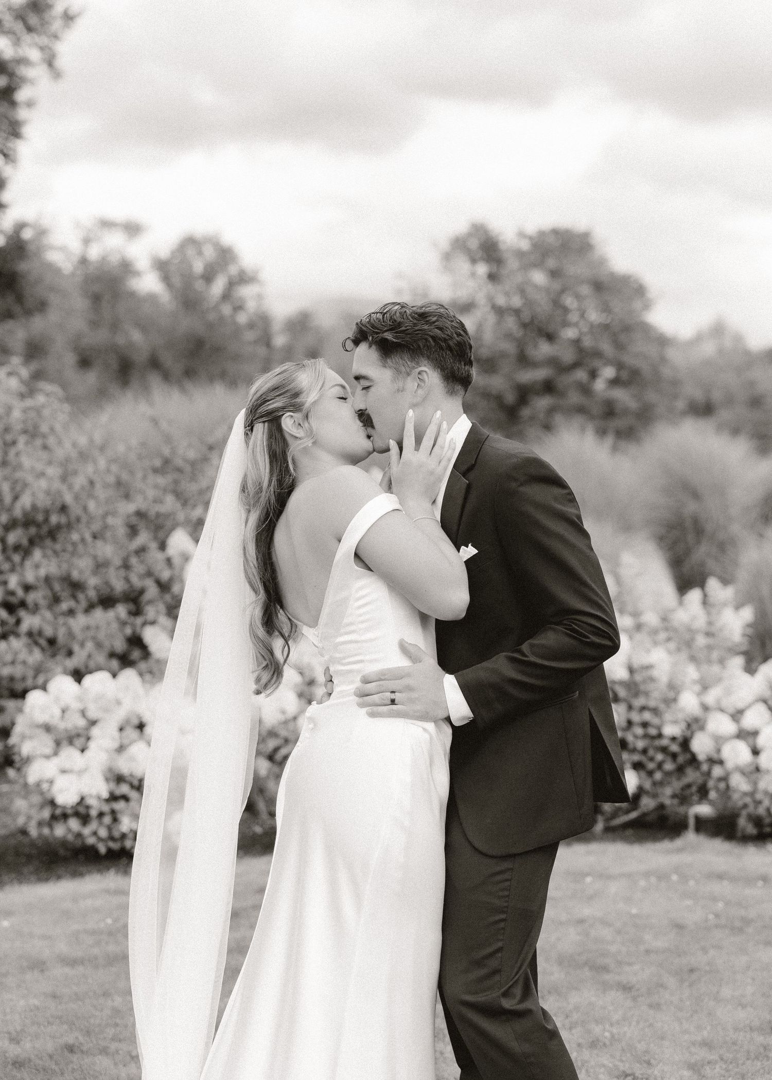 Black and white photo of a bride and groom sharing a romantic kiss outdoors with lush garden blooms in the background.