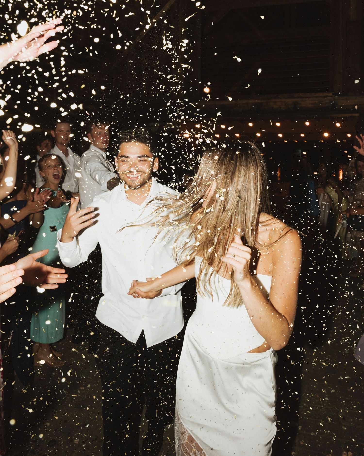 Couple in white attire dancing joyfully amid confetti shower at outdoor wedding celebration with guests cheering.