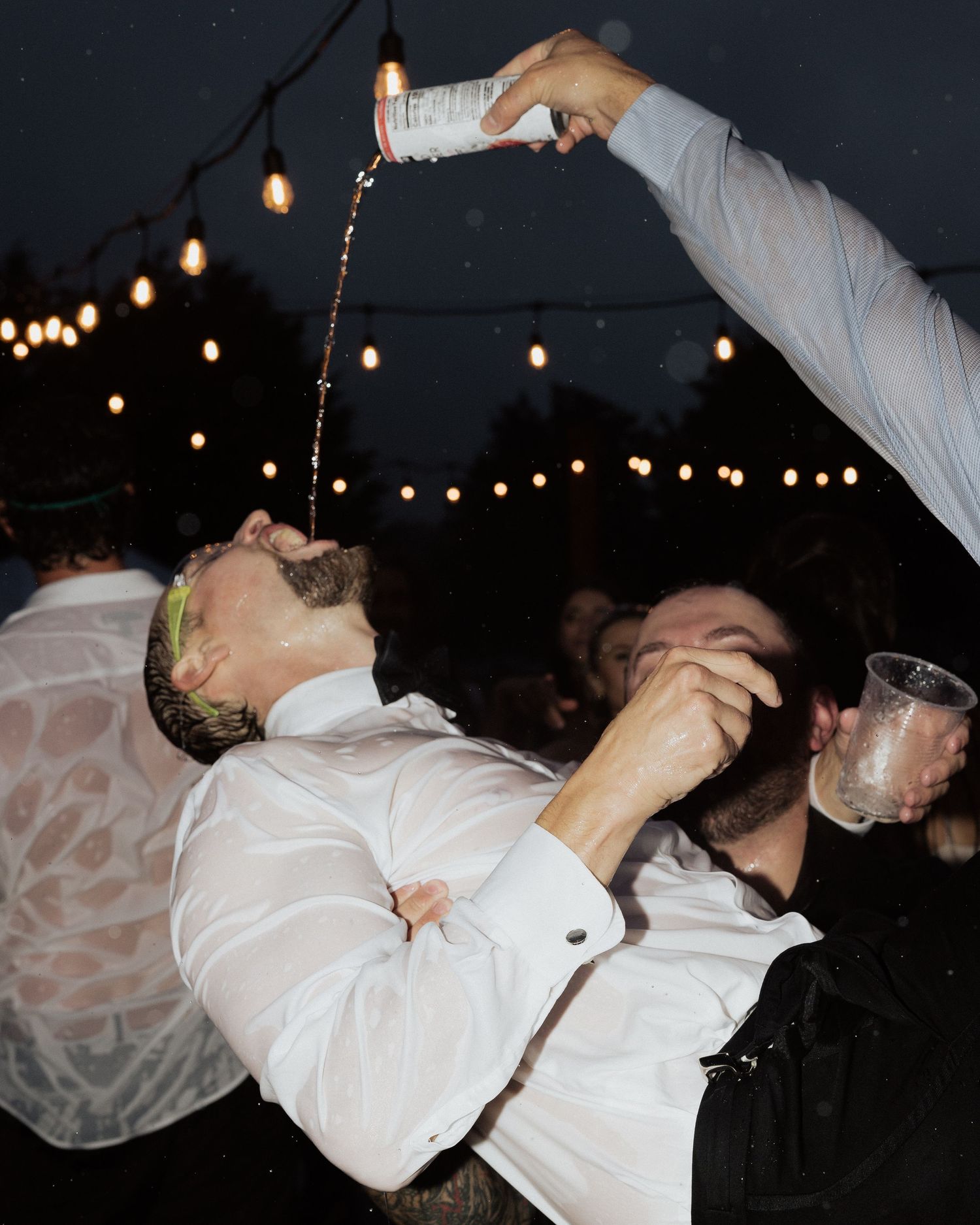 Someone pouring a drink into another person's mouth at an outdoor party under string lights.