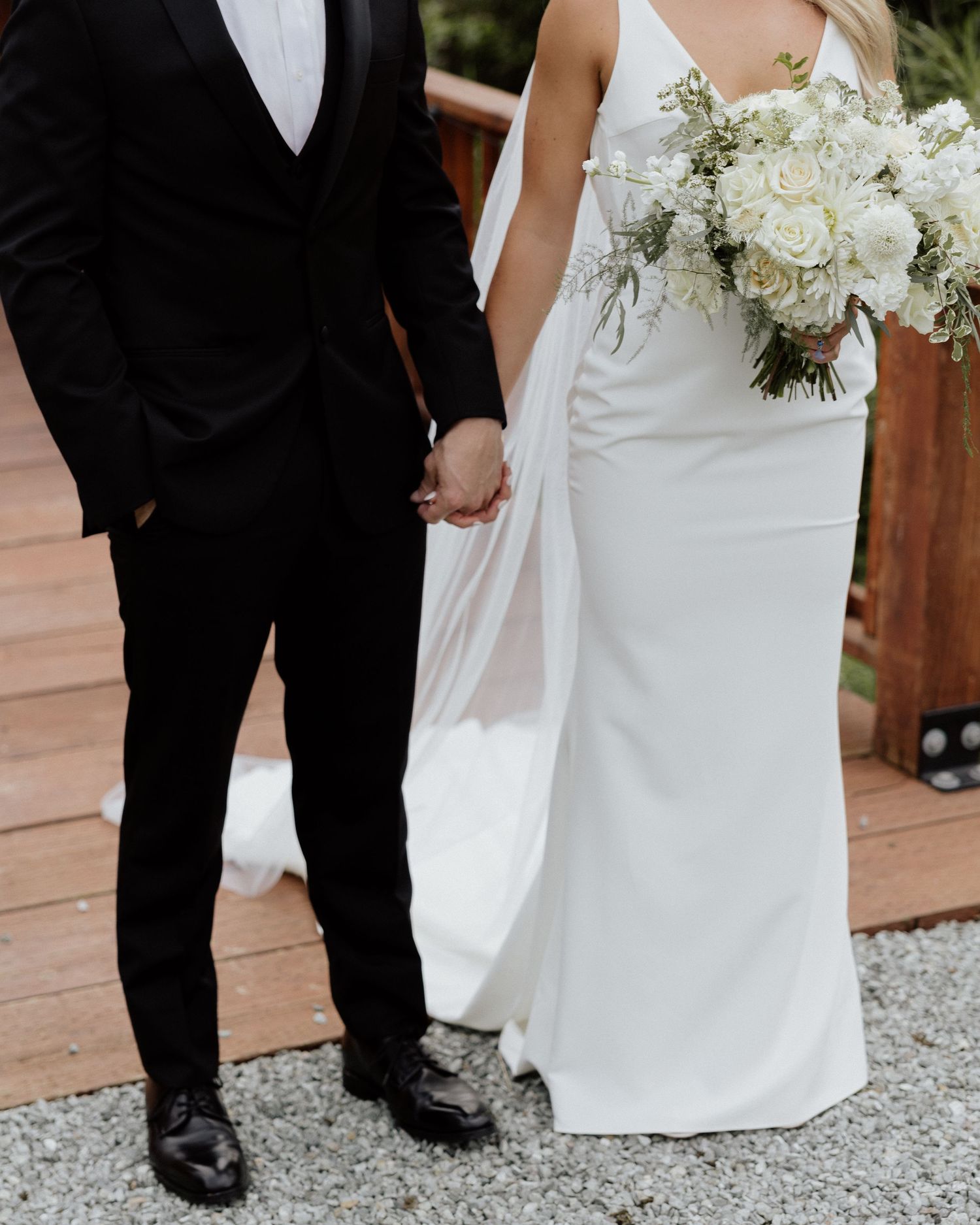 Elegant couple holding hands showing wedding attire details and white bouquet.
