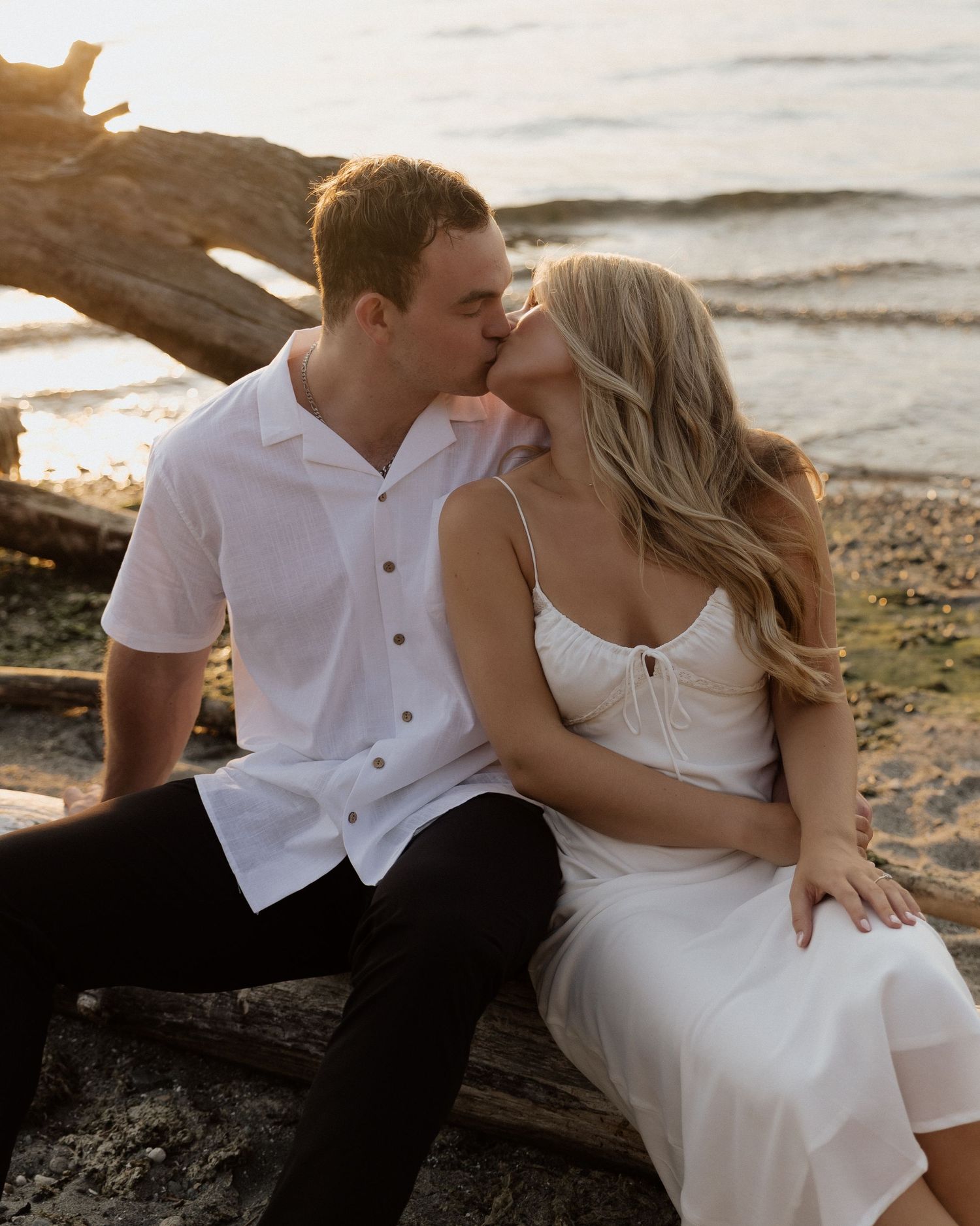 A couple shares an intimate moment while sitting on driftwood at a beach during sunset.