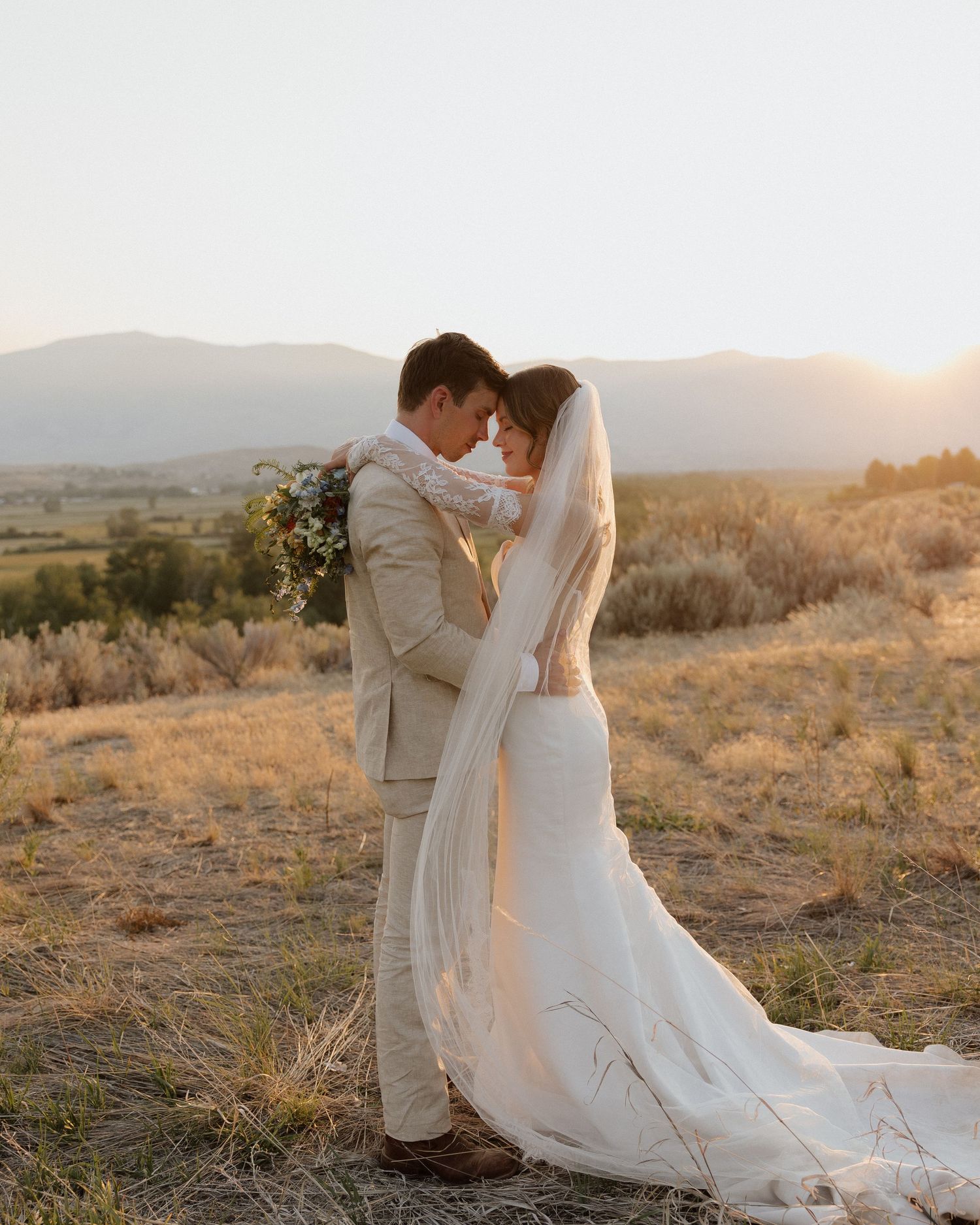 Bride and groom share tender moment on hillside with flowing veil and desert mountain backdrop.