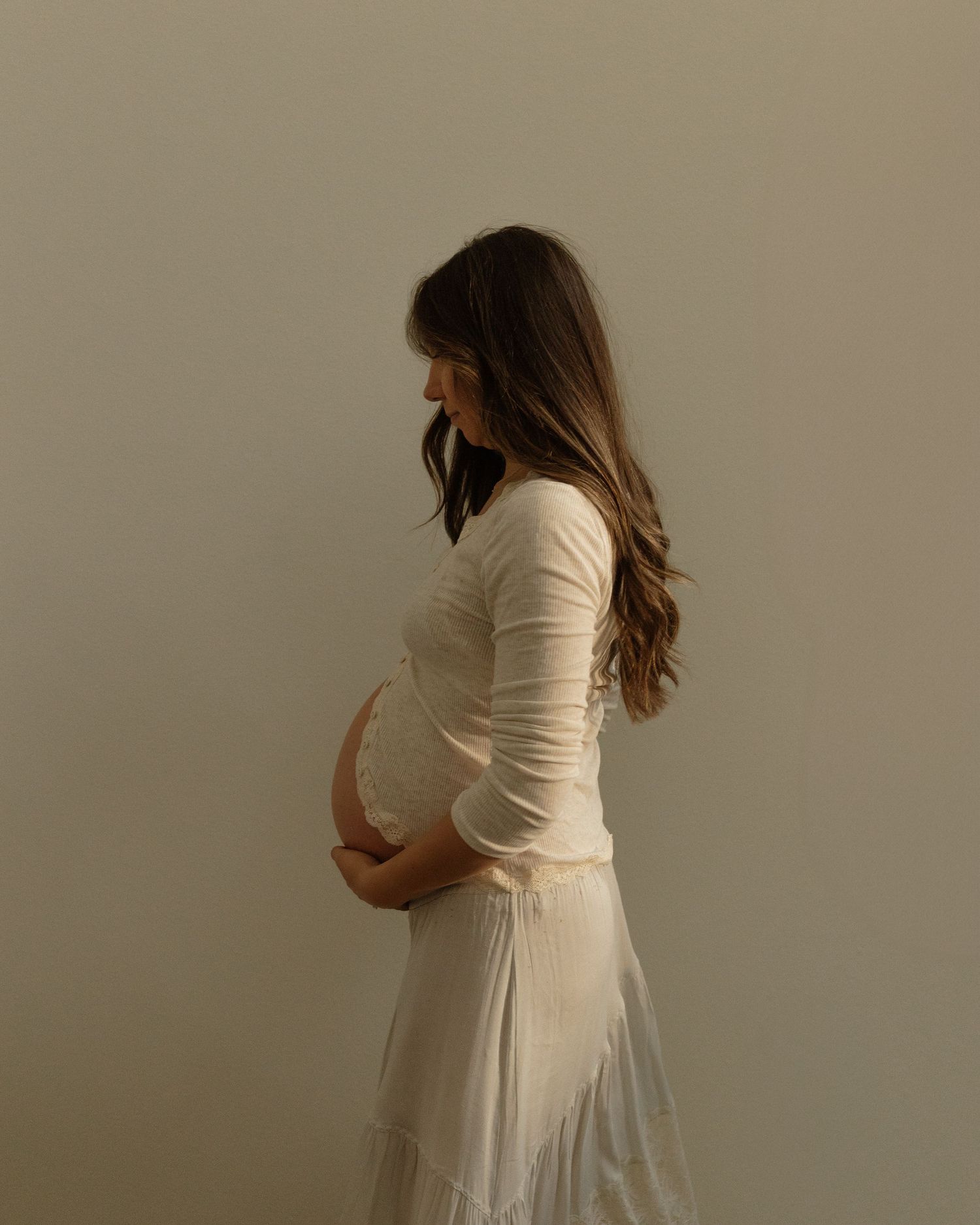 An elegant side profile maternity photo of a woman in white holding her baby bump.