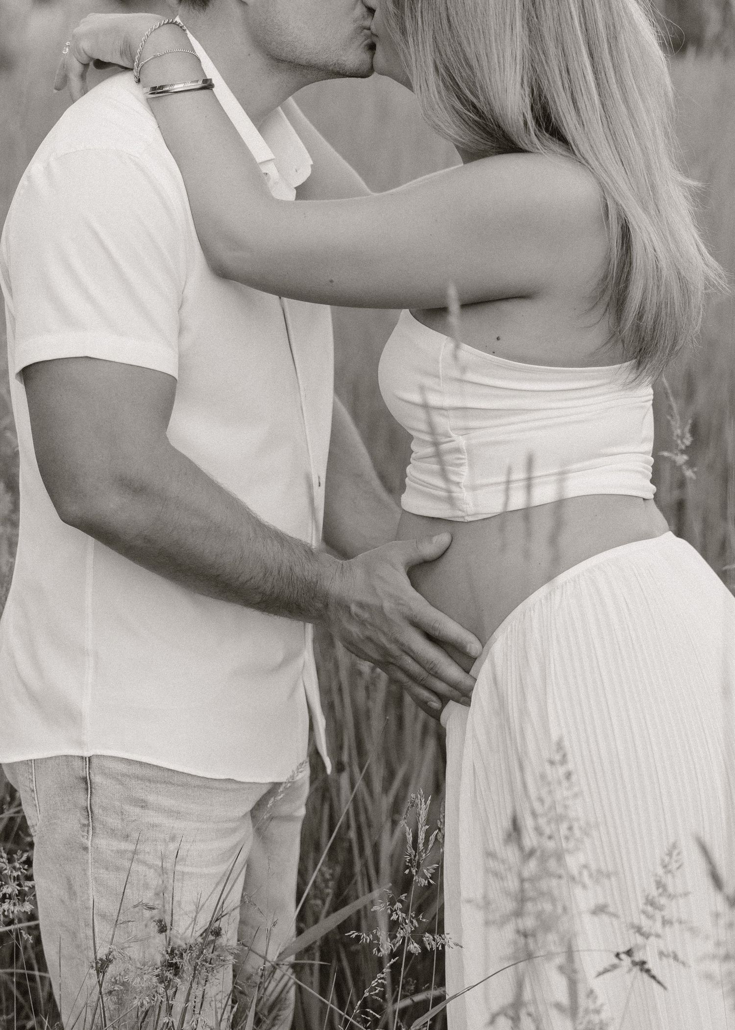 Black and white photo of couple embracing in a field, man tenderly holding pregnant woman's baby bump.