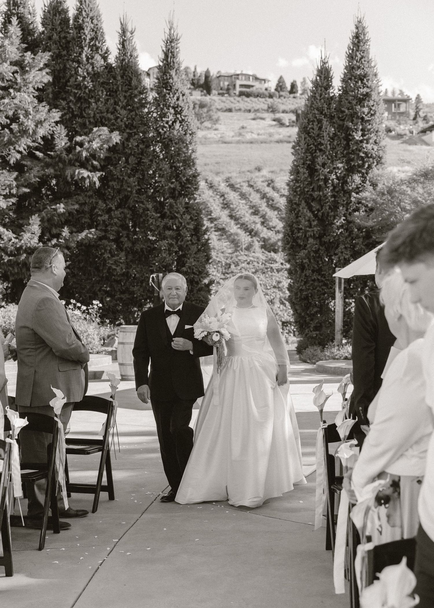 Black and white photo of a bride walking down the aisle with her father at an outdoor vineyard wedding ceremony.