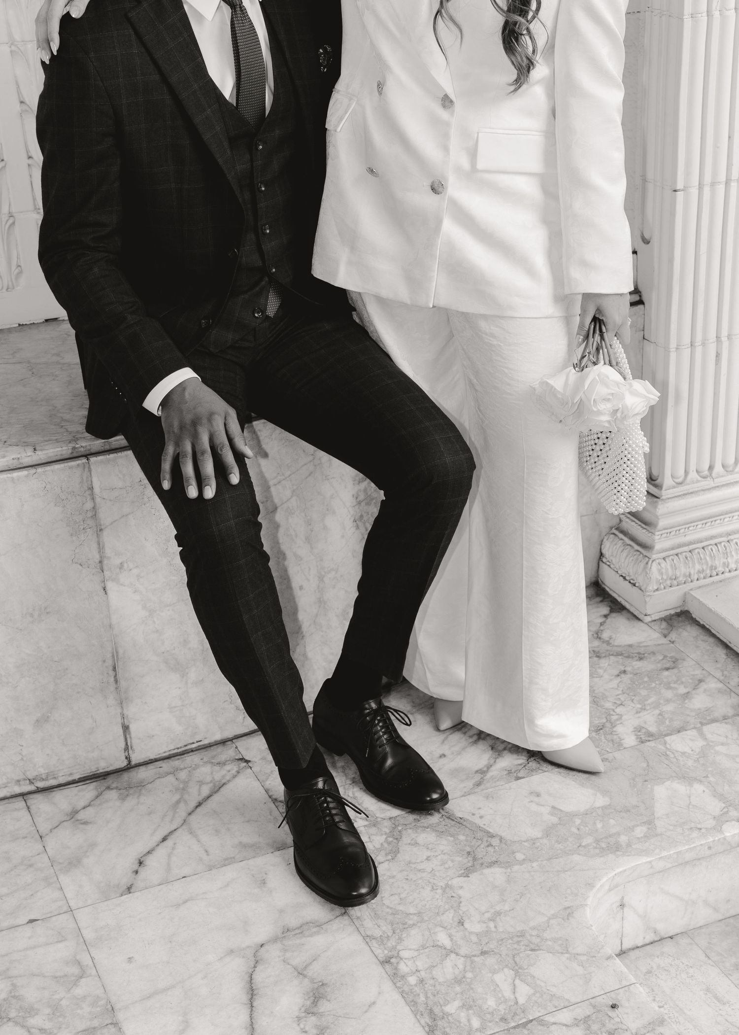 Black and white photo of a bride in white suit and groom in dark suit sitting on marble steps at wedding.