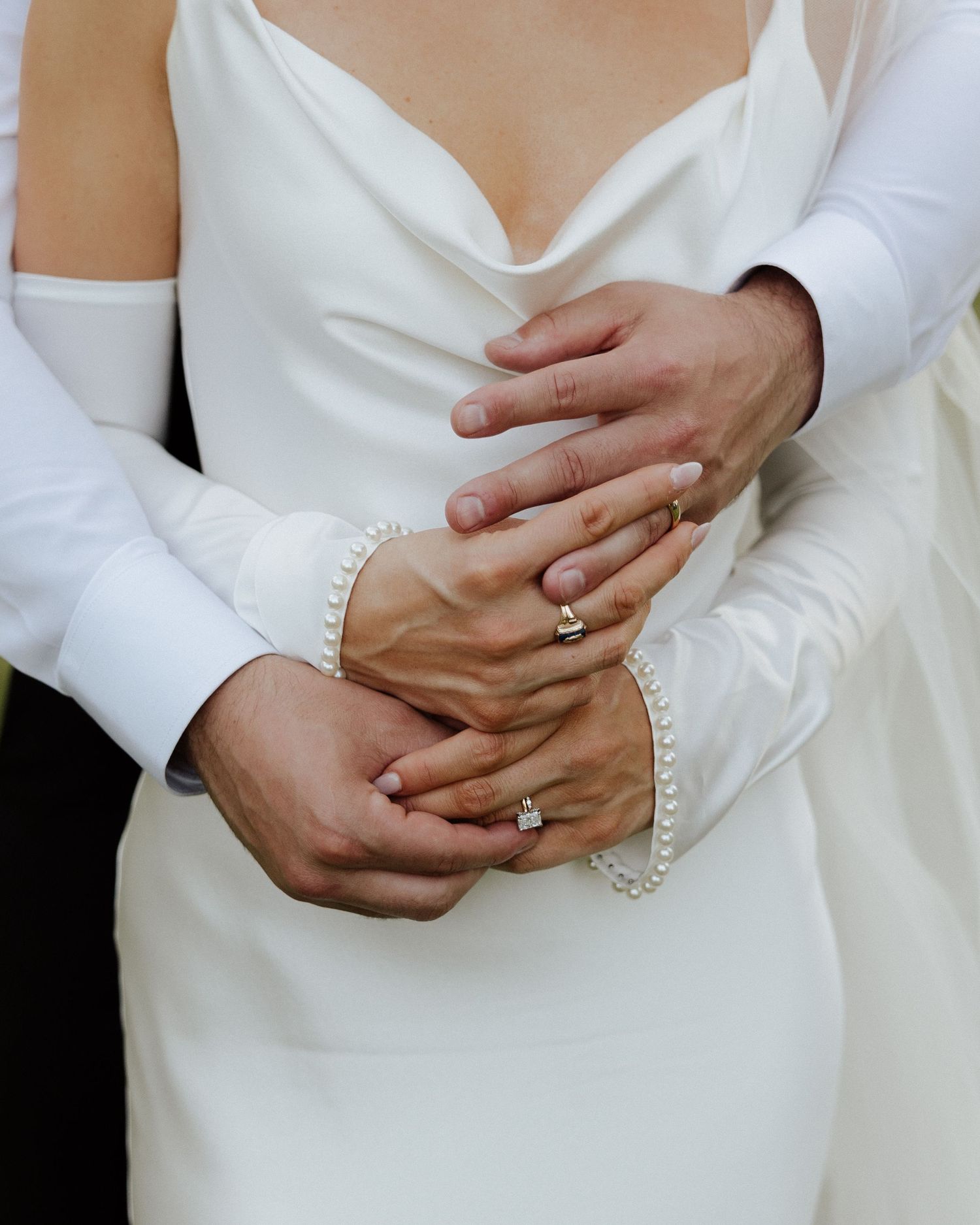Close-up of newlyweds hands intertwined, bride wearing diamond ring and pearl bracelet in white wedding dress.