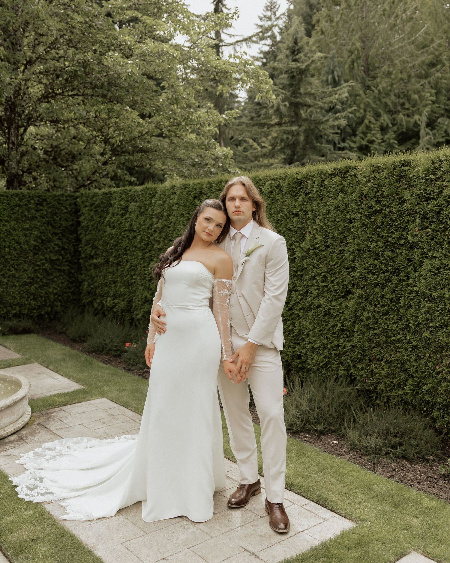 Bride and groom posing together in lush garden, she in white gown, he in cream suit.