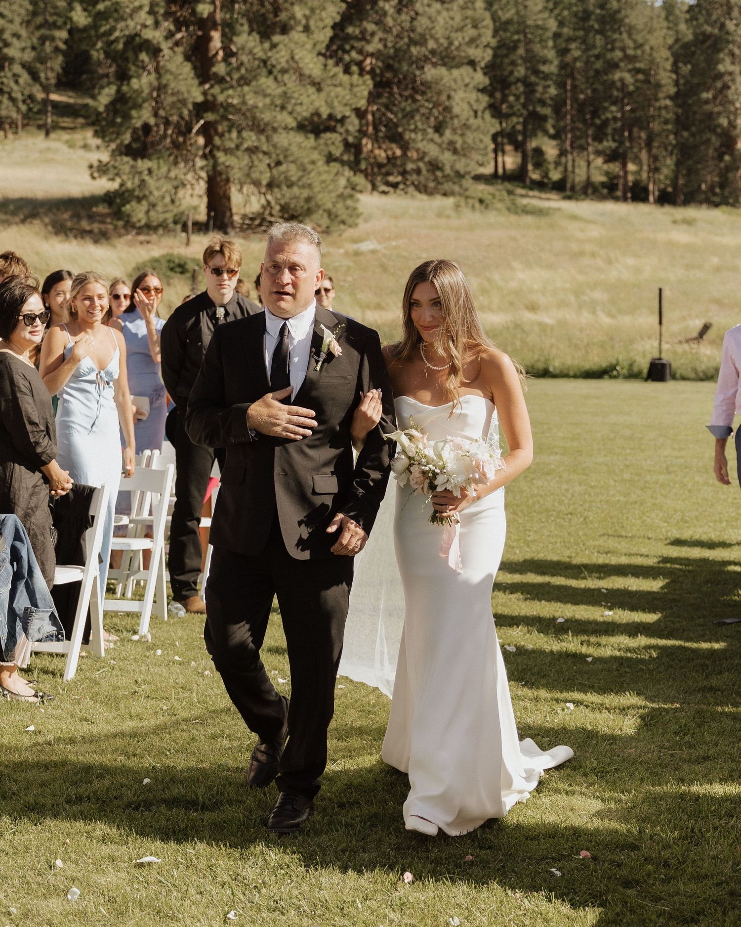 Bride in white gown walks down outdoor aisle with father during sunny wedding ceremony on green lawn.