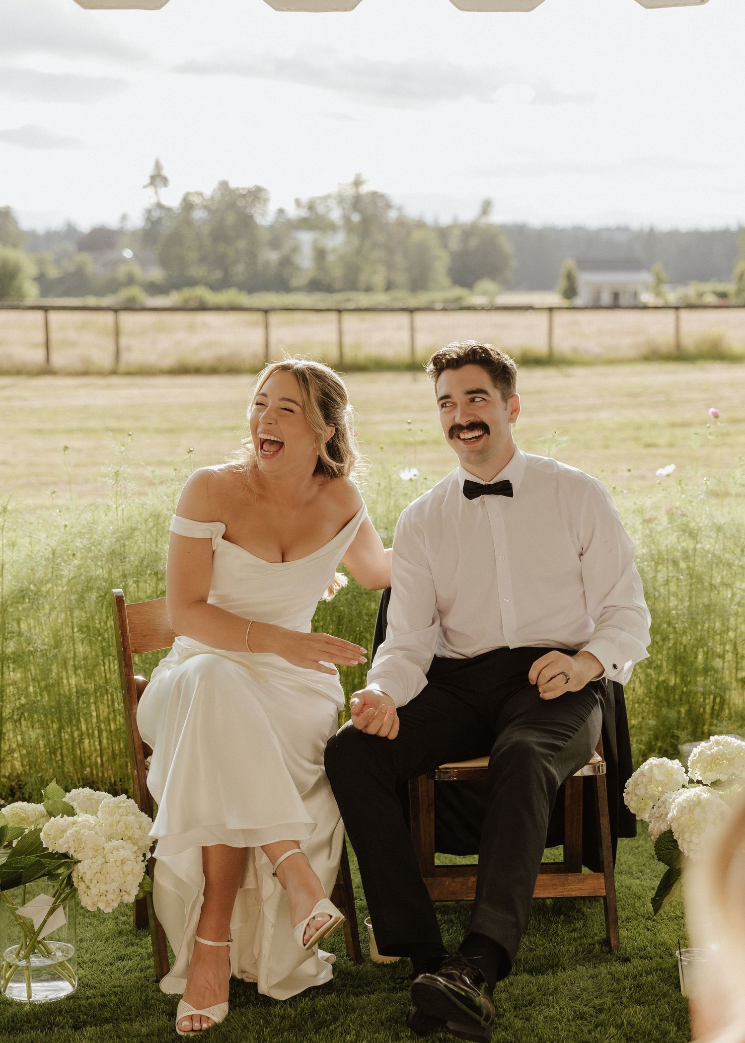 Laughing bride and groom with mustache sit together on wooden chairs at outdoor summer wedding reception.
