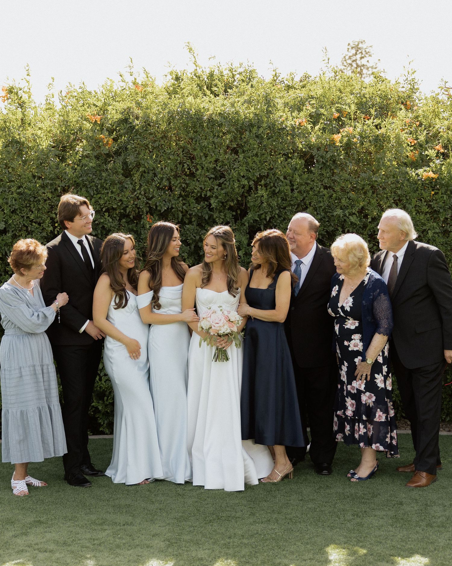 Wedding party group photo with bride in white gown, bridesmaids in blue dresses, and family members outdoors.